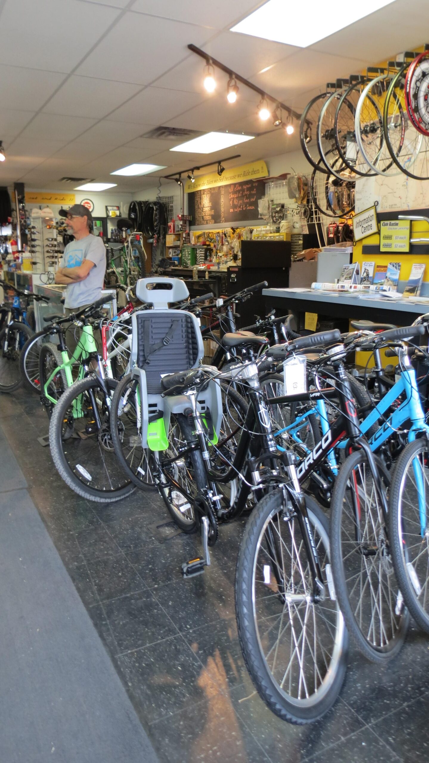 An interior view of a bicycle shop featuring various bicycles on display, including mountain bikes and a bike with a child seat attached. A man stands in the background, and there are shelves with bike accessories and a chalkboard listing bike maintenance tips. The shop has bright lighting and a welcoming atmosphere.