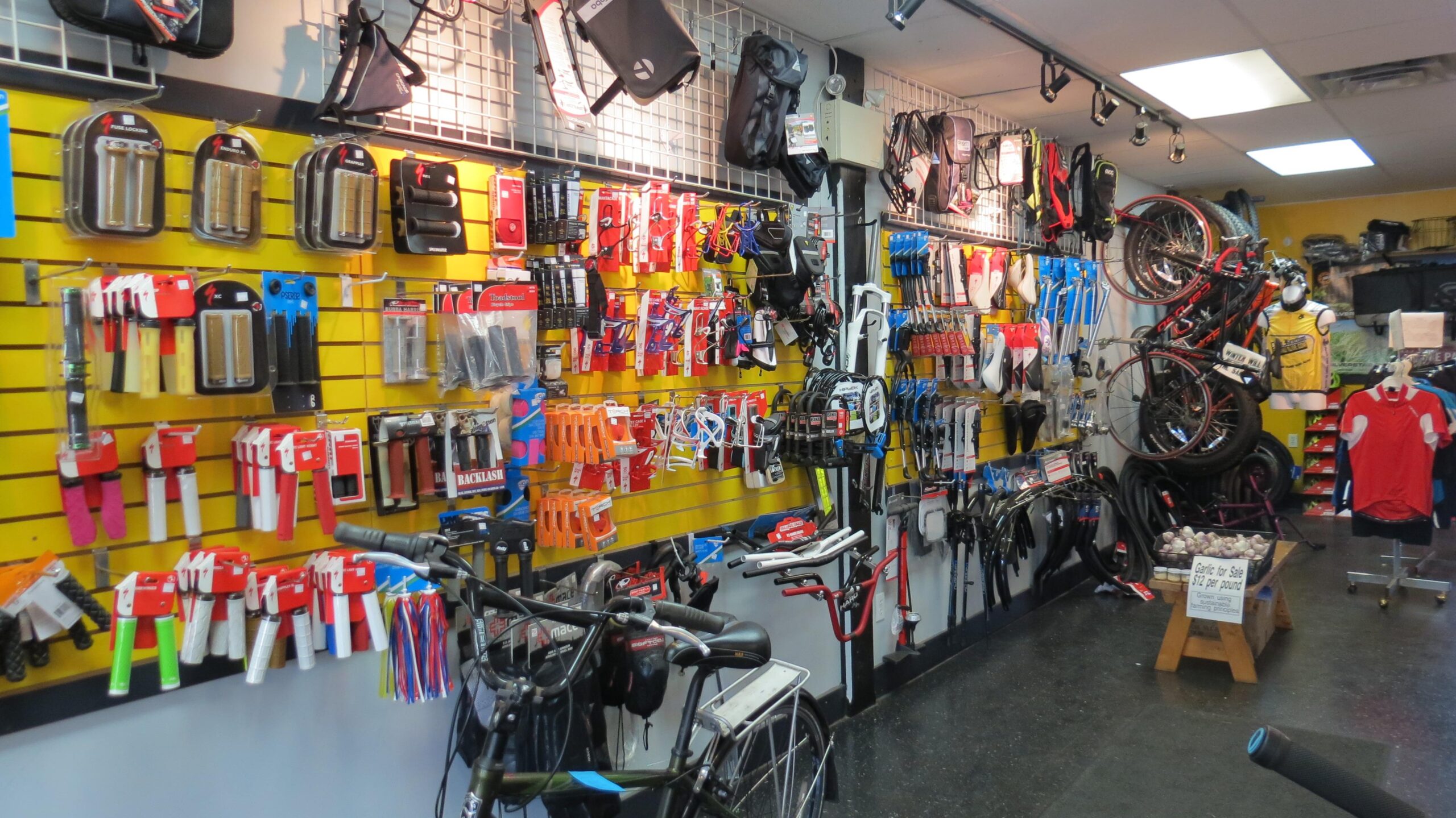 Image of a bike shop interior showcasing various bicycle accessories and parts. The walls are lined with tools, grips, and components on pegboards, while bicycles are displayed prominently. A small table features garlic for sale, and a red cycling jersey is visible hanging in the background. The shop has a bright and organized atmosphere, highlighting the range of products available for cycling enthusiasts.