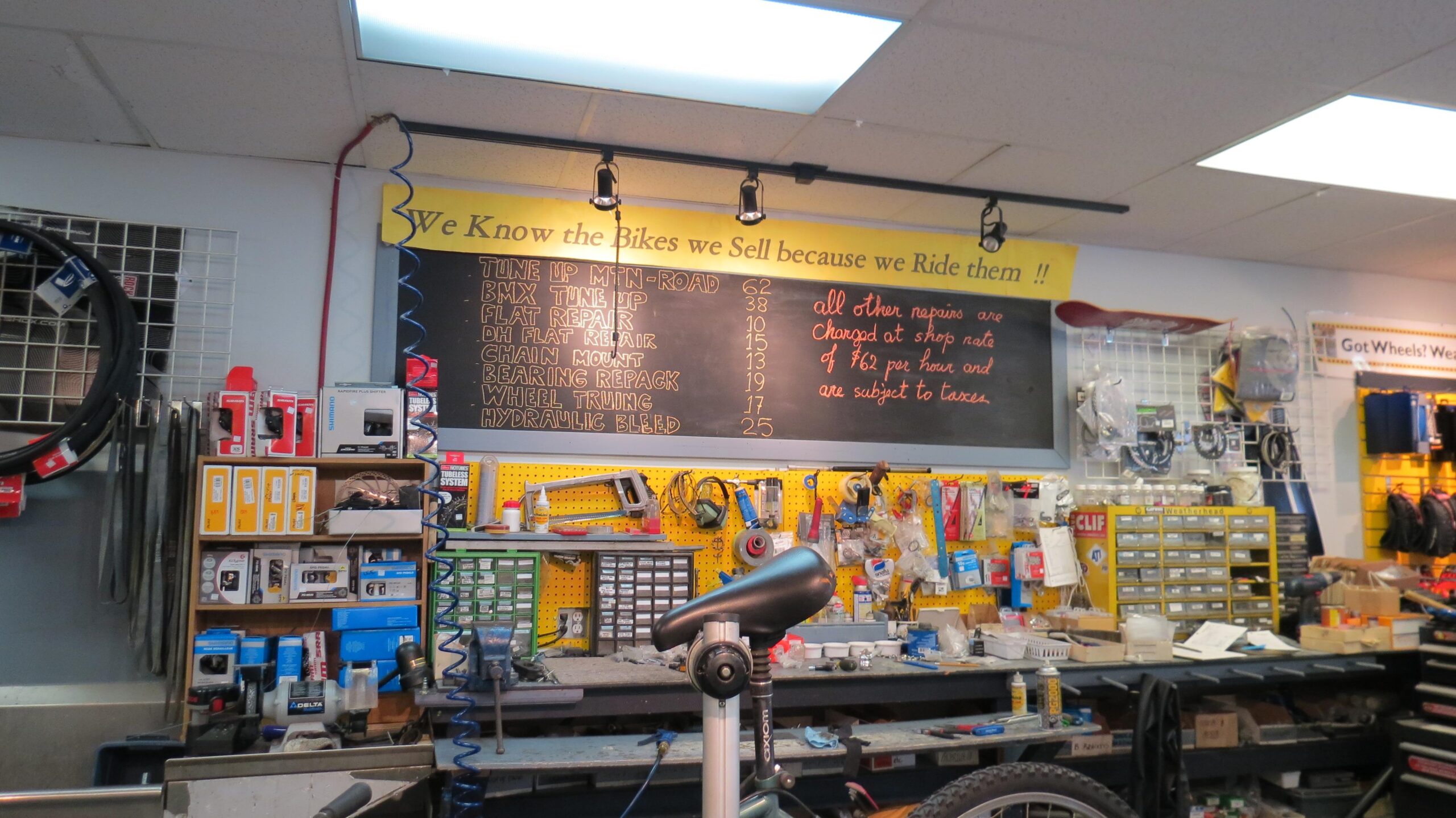 A bicycle repair shop interior featuring a black chalkboard with a list of repair prices, tools, and bike parts organized on shelves and workbenches. The text on the chalkboard details various services such as "Tune Up," "Flat Repair," and "Wheel Truing," along with their respective prices. The shop is cluttered with a variety of bicycle parts, tools, and accessories, indicating a busy and functional workspace. A sign above the chalkboard reads, "We Know the Bikes we Sell because we Ride them !!"