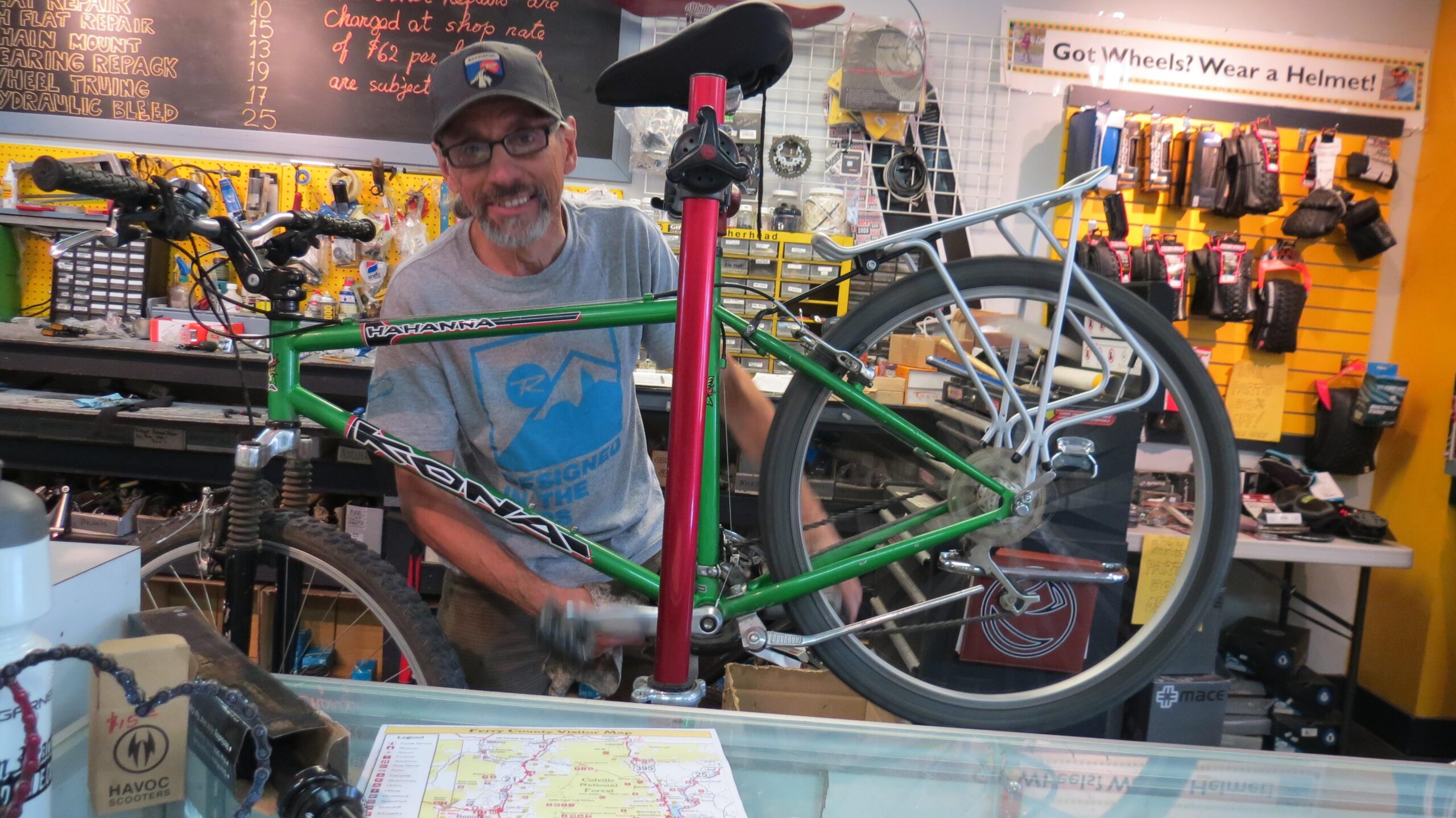 A smiling mechanic working on a green mountain bike in a bicycle repair shop, with various tools and bike parts visible in the background. A sign on the wall reminds customers to wear helmets while cycling.