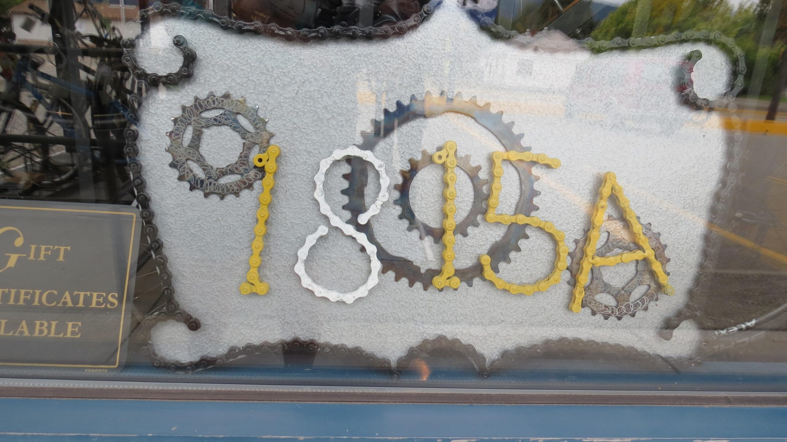 A decorative window display featuring the numbers "1895A" created from colored bicycle chain links and gears on a textured background. In the lower left corner, there is a sign that reads "Gift Certificates Available."