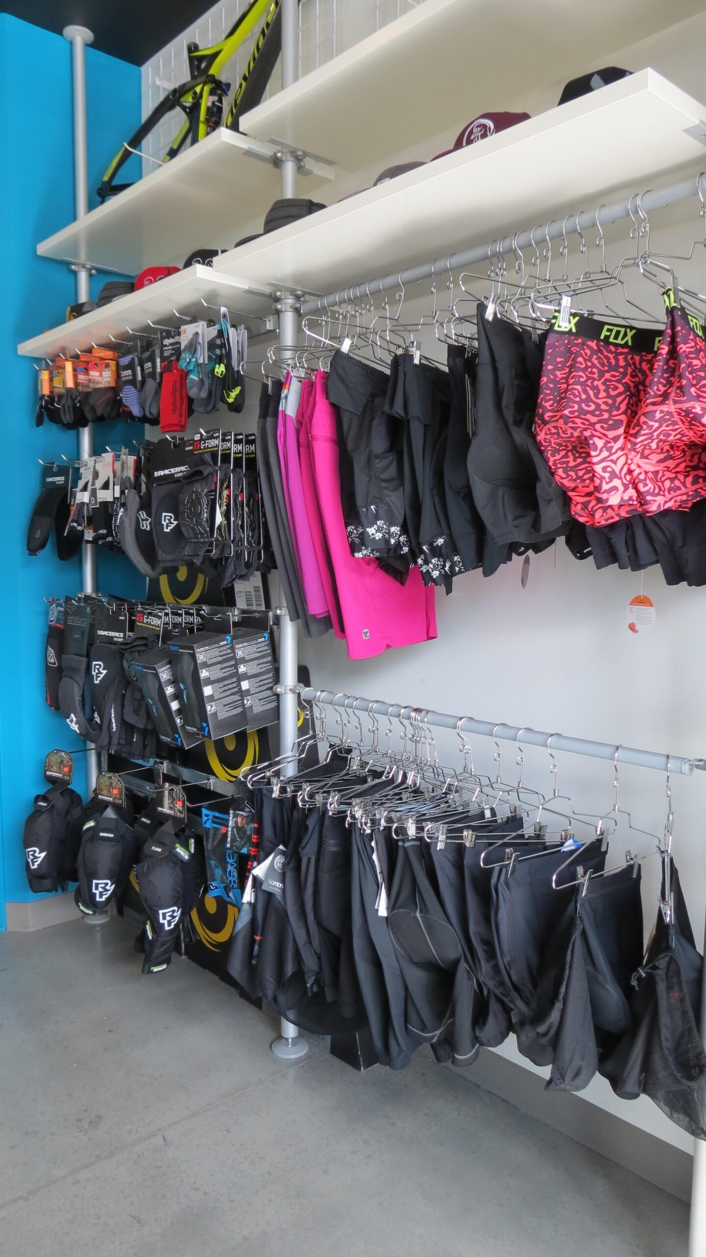 A display wall in a retail store featuring various cycling apparel and accessories. The rack includes black shorts, gloves, protective gear, and some colorful items like bright pink shorts with a patterned design. Above, hats and additional gear are displayed on shelves, with a bicycle hanging in the background against a blue wall. The store has a clean and organized appearance, showcasing a variety of biking products.