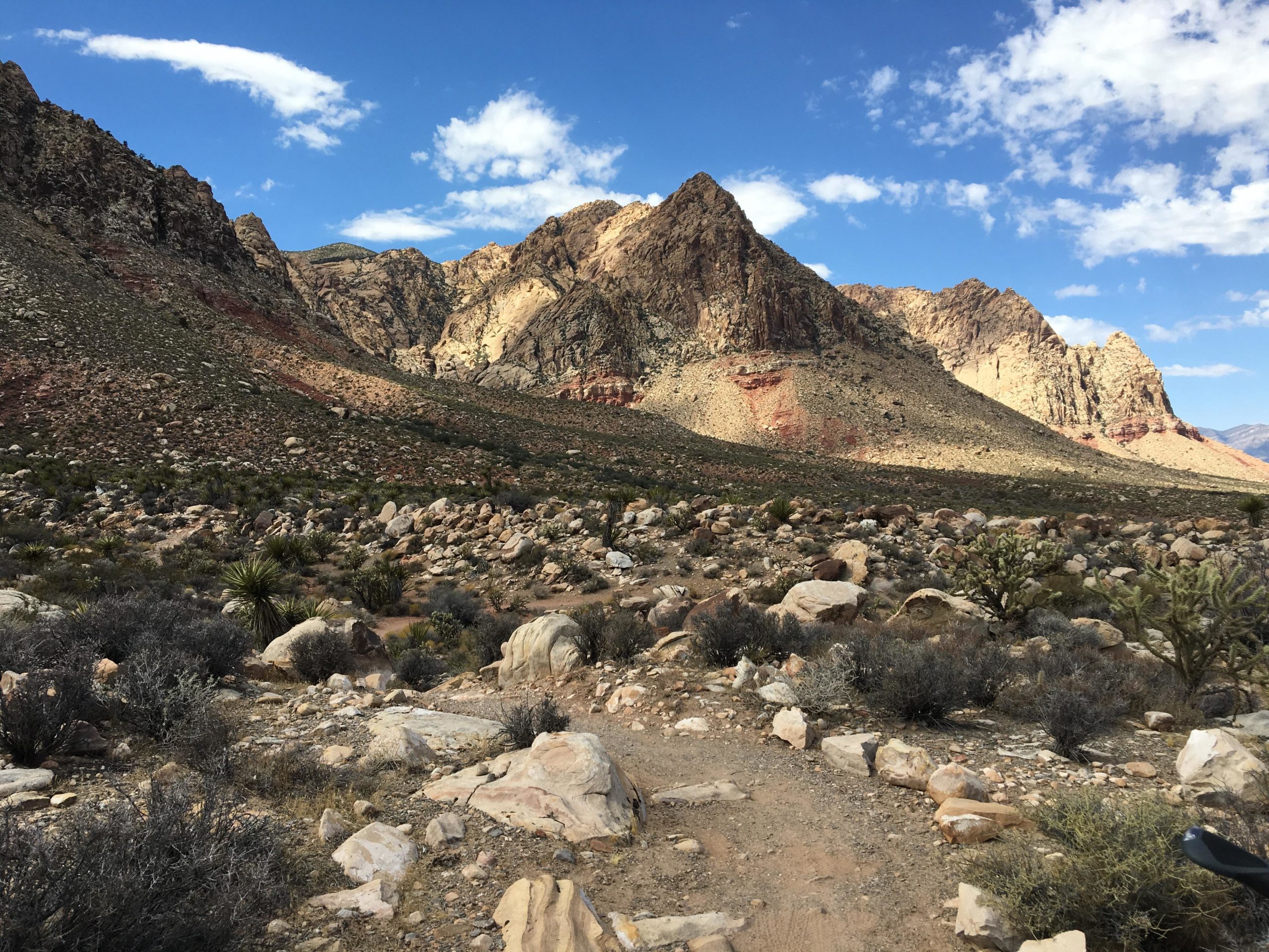 A rocky desert landscape featuring a winding dirt path, scattered boulders, and sparse vegetation, framed by rugged mountains under a partly cloudy blue sky. Cottonwood Valley North mountain bike trail.