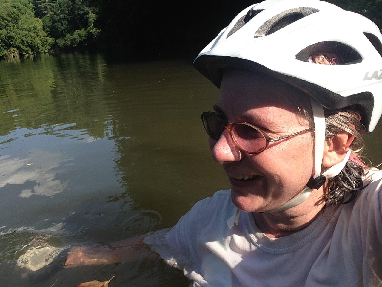 A person wearing a helmet and sunglasses smiles while sitting in a body of water. The background features lush green trees reflecting in the water, under a clear blue sky. Overmountain Victory Trail mountain bike trail.