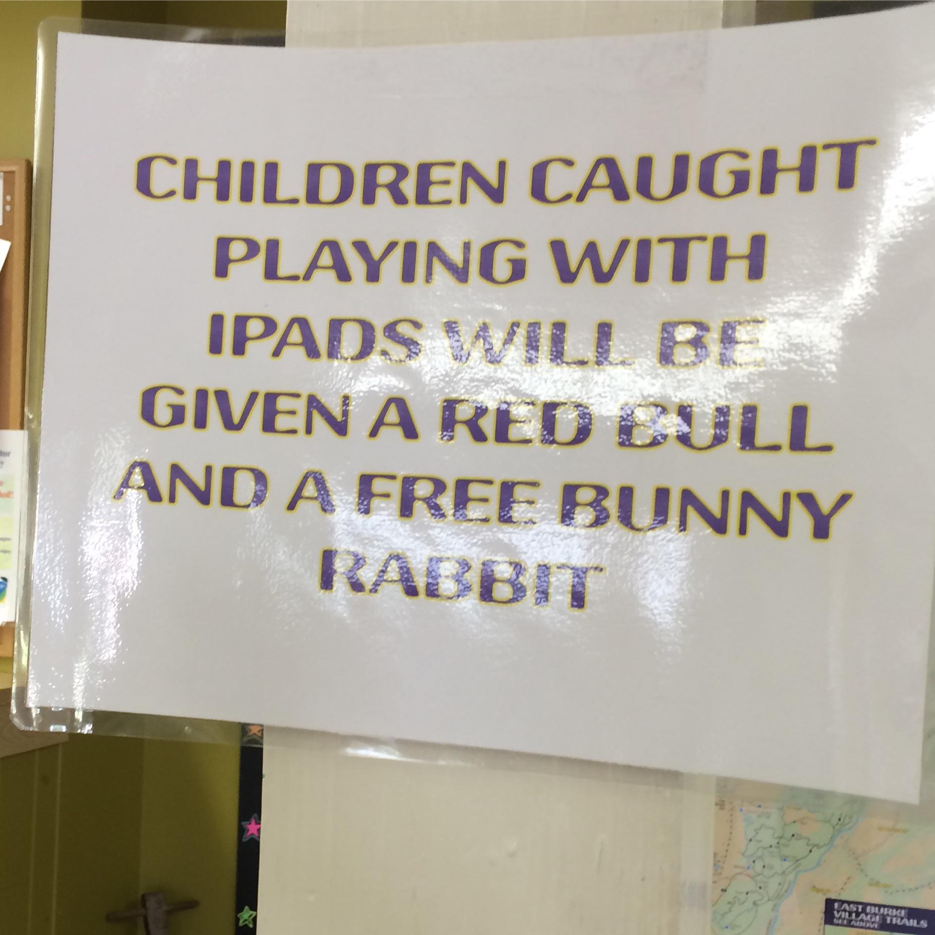 A humorous sign posted on a wall that reads: "Children caught playing with iPads will be given a Red Bull and a free bunny rabbit." The text is bold and colorful, highlighting a playful warning. Kingdom Trails mountain bike trail.