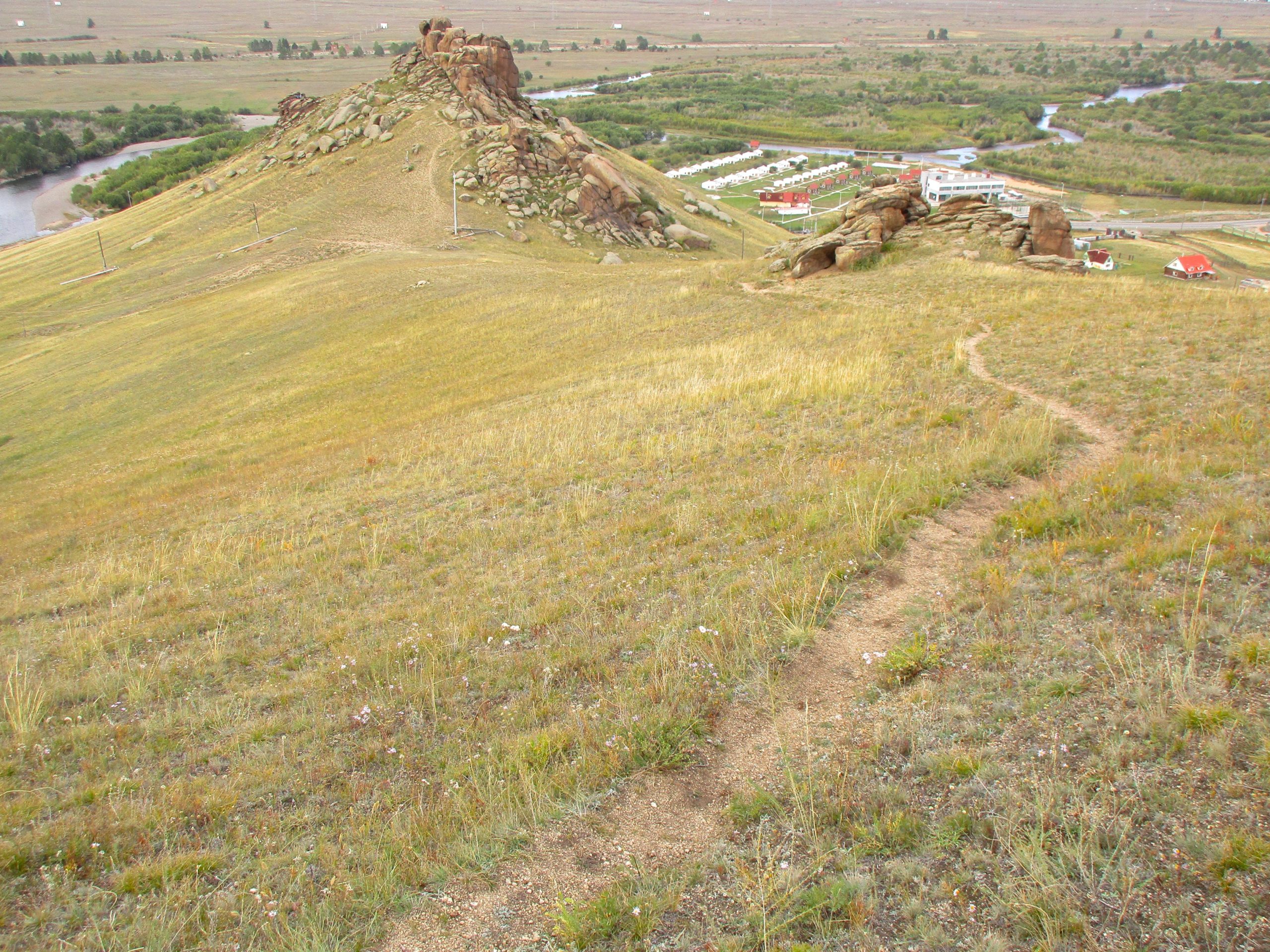 A scenic view of a grassy hillside leading to rocky formations, with a winding dirt path. In the background, a river flows through a green valley, and a series of buildings with red roofs are visible near the water. The landscape features a mix of dry grass and scattered wildflowers under an expansive sky. The Beast mountain bike trail.
