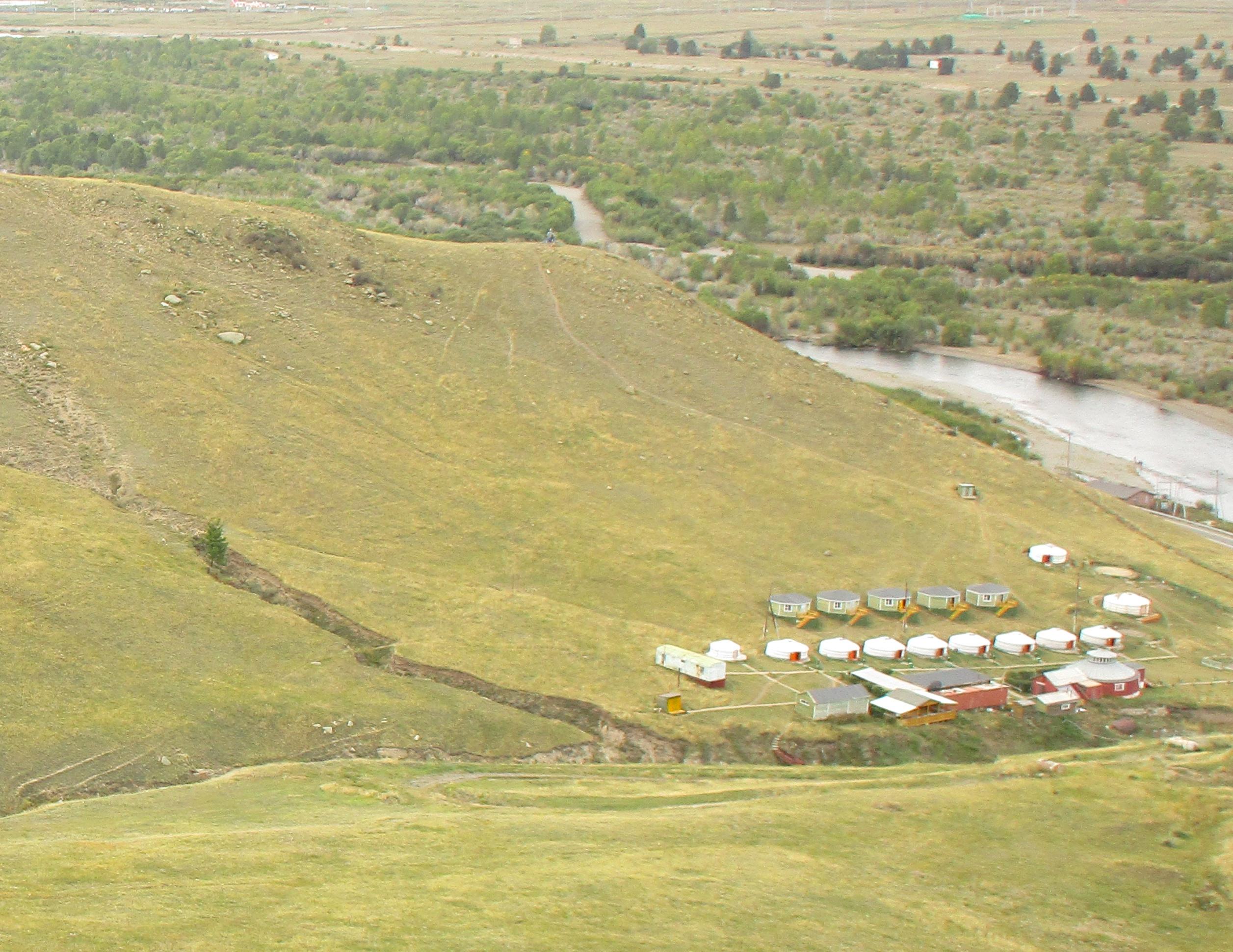 Aerial view of a grassy hillside with several buildings at the base, including a row of colorful cabins or yurts, and a nearby winding river surrounded by trees. The landscape features a mix of vibrant green vegetation and gold-hued grassy hills. The Beast mountain bike trail.