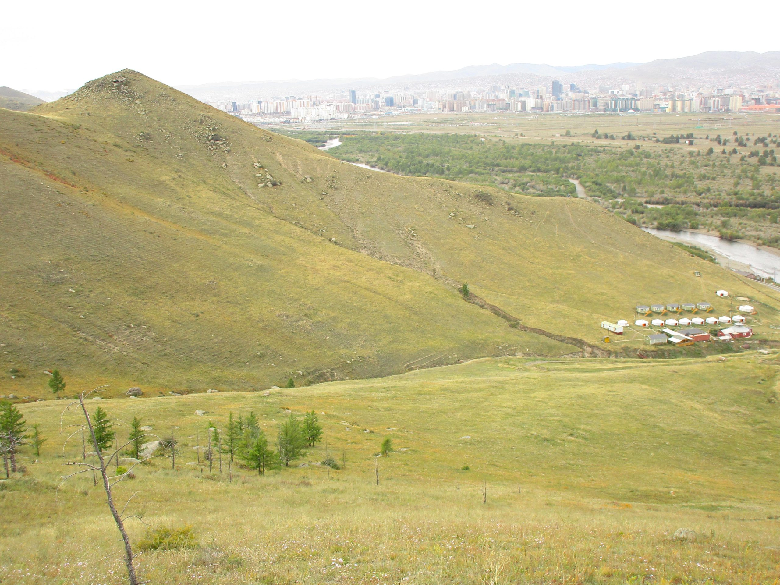 A panoramic view of a grassy hillside leading down to a river, with a small cluster of traditional yurts on the right. In the background, a city skyline is visible against a backdrop of distant mountains under a cloudy sky. The Beast mountain bike trail.