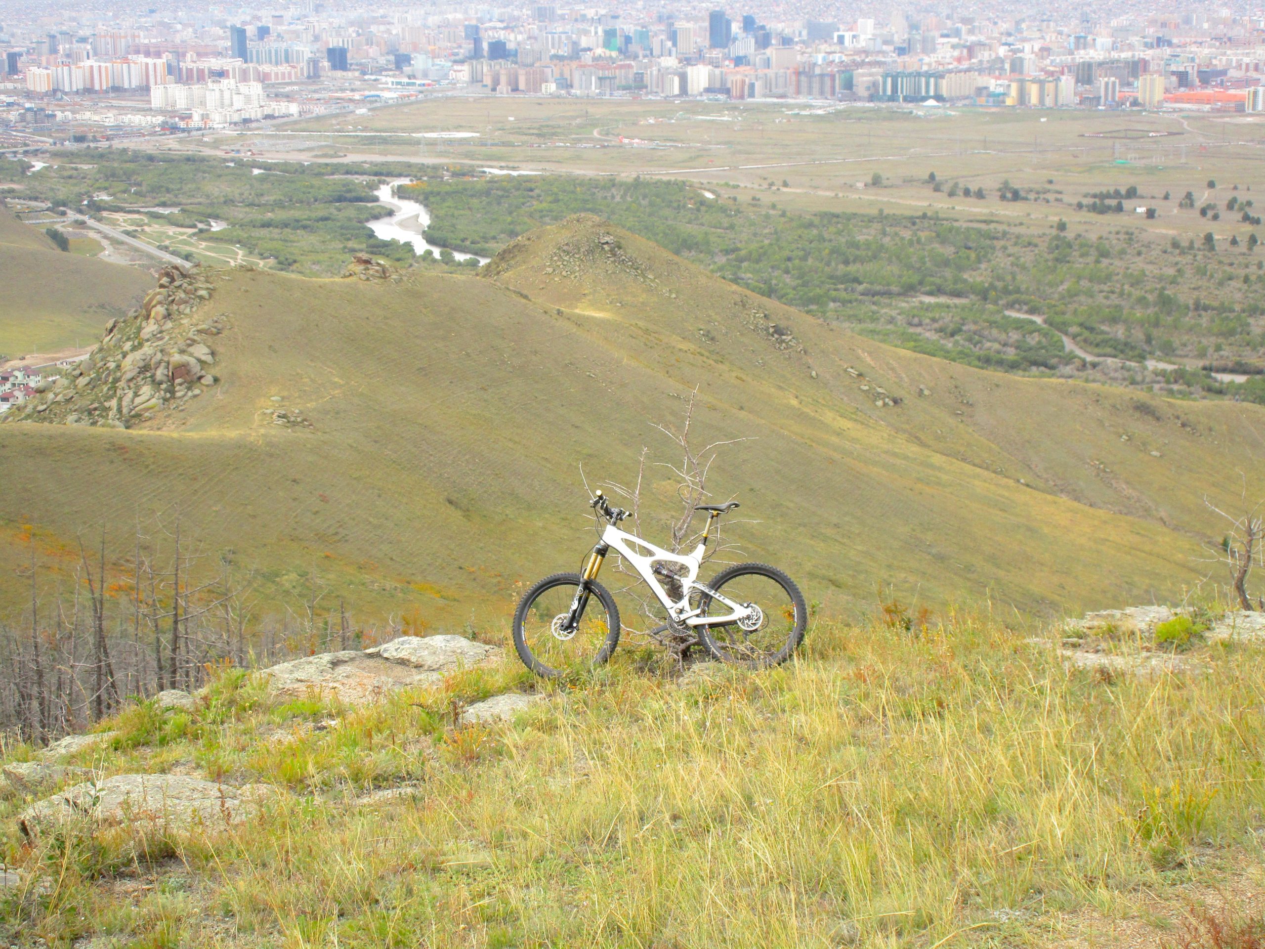A mountain bike resting on a rocky outcrop, overlooking a vast landscape of rolling hills, greenery, and a distant city skyline. The scene captures a blend of nature and urban development, with a river winding through the valley below. The Beast mountain bike trail.