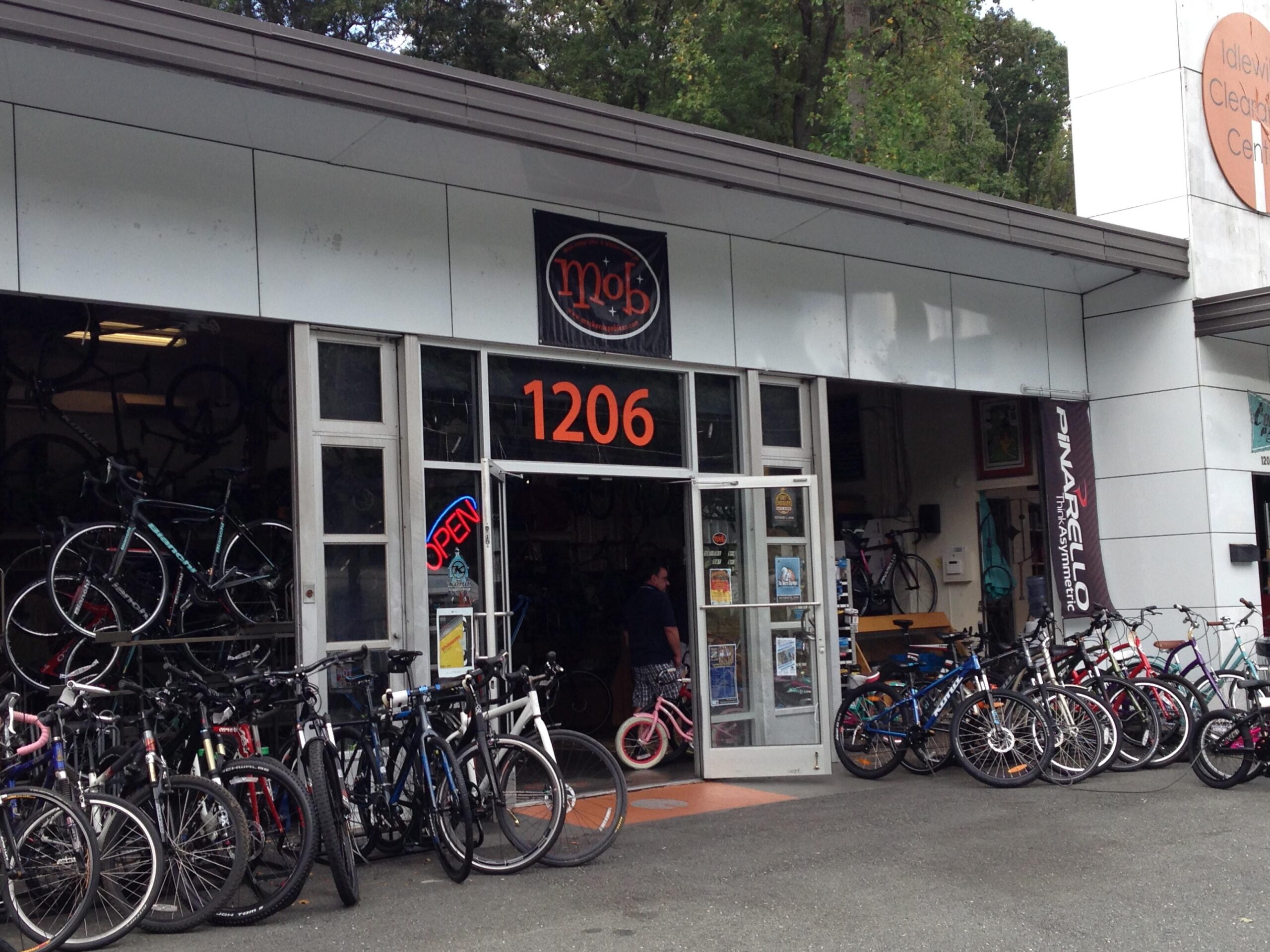 Front view of a bicycle shop featuring a variety of bicycles displayed outside. The store has a large glass entrance with "OPEN" signage and the address "1206" prominently displayed. Various styles of bikes, including road and mountain bikes, are neatly arranged in front of the shop. A person is visible near the entrance, and several bicycles can be seen inside through the glass doors.