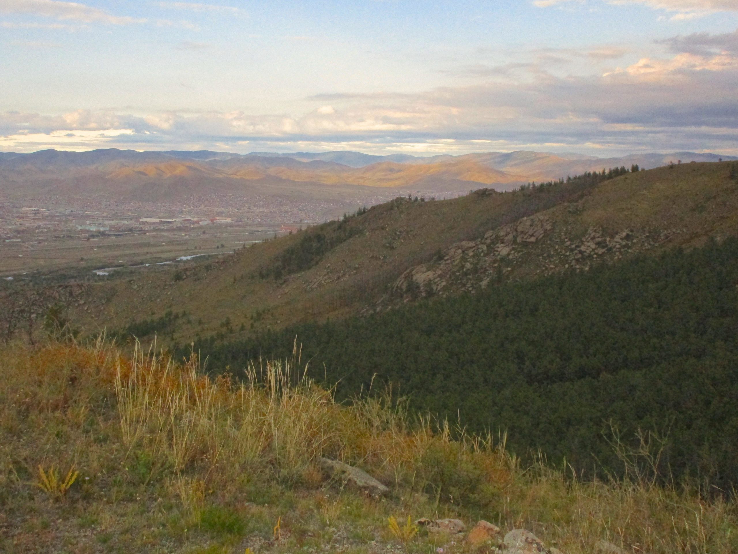 A panoramic view of rolling hills and distant mountains under a cloudy sky. The foreground features a patch of green vegetation and rocky terrain, while a valley below reveals a scenic landscape with a small town and fields. The light of the setting sun casts warm tones across the hills. The Beast mountain bike trail.