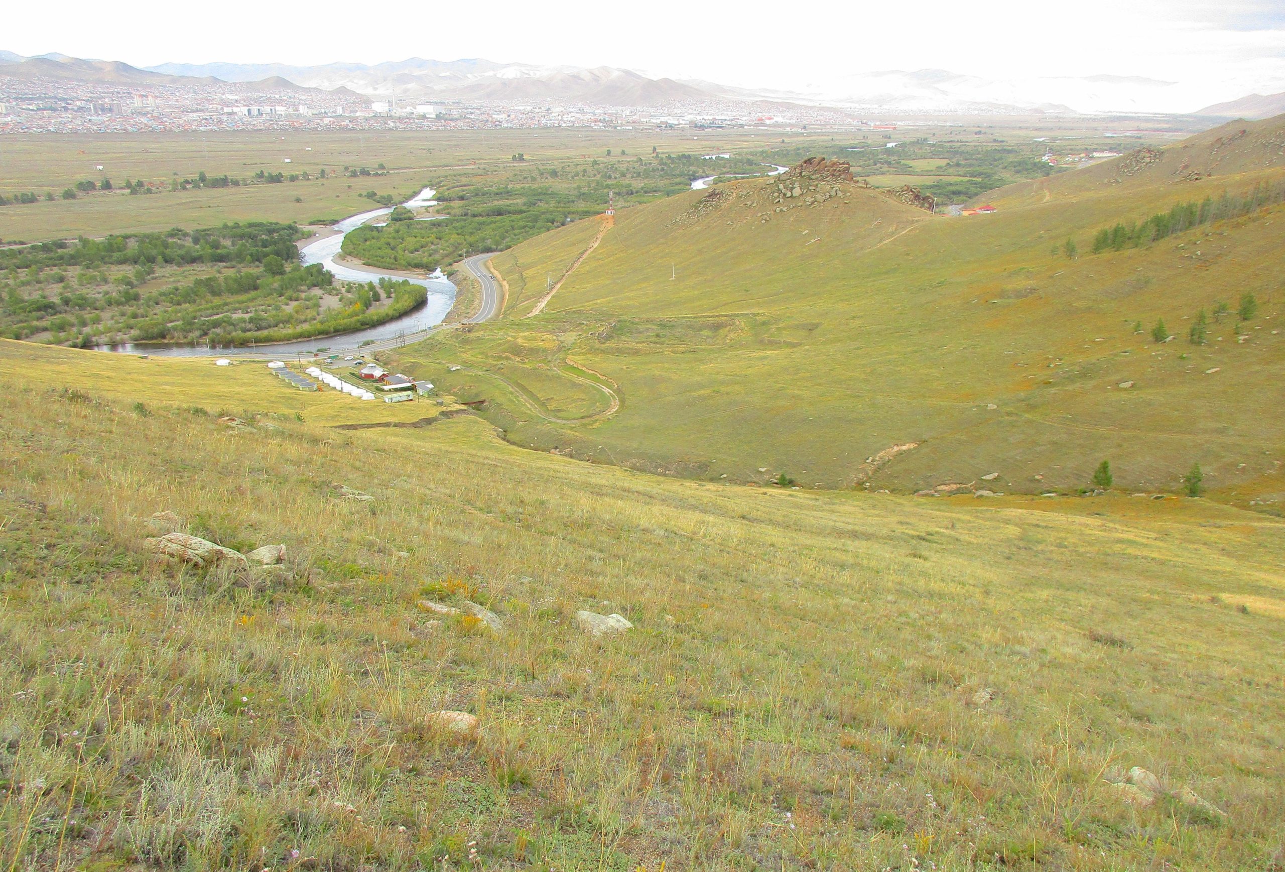 A panoramic view of a valley featuring a winding river surrounded by greenery and rolling hills. The foreground shows a grassy slope with scattered rocks, while the background reveals a distant city skyline and mountains under a cloudy sky. The Beast mountain bike trail.