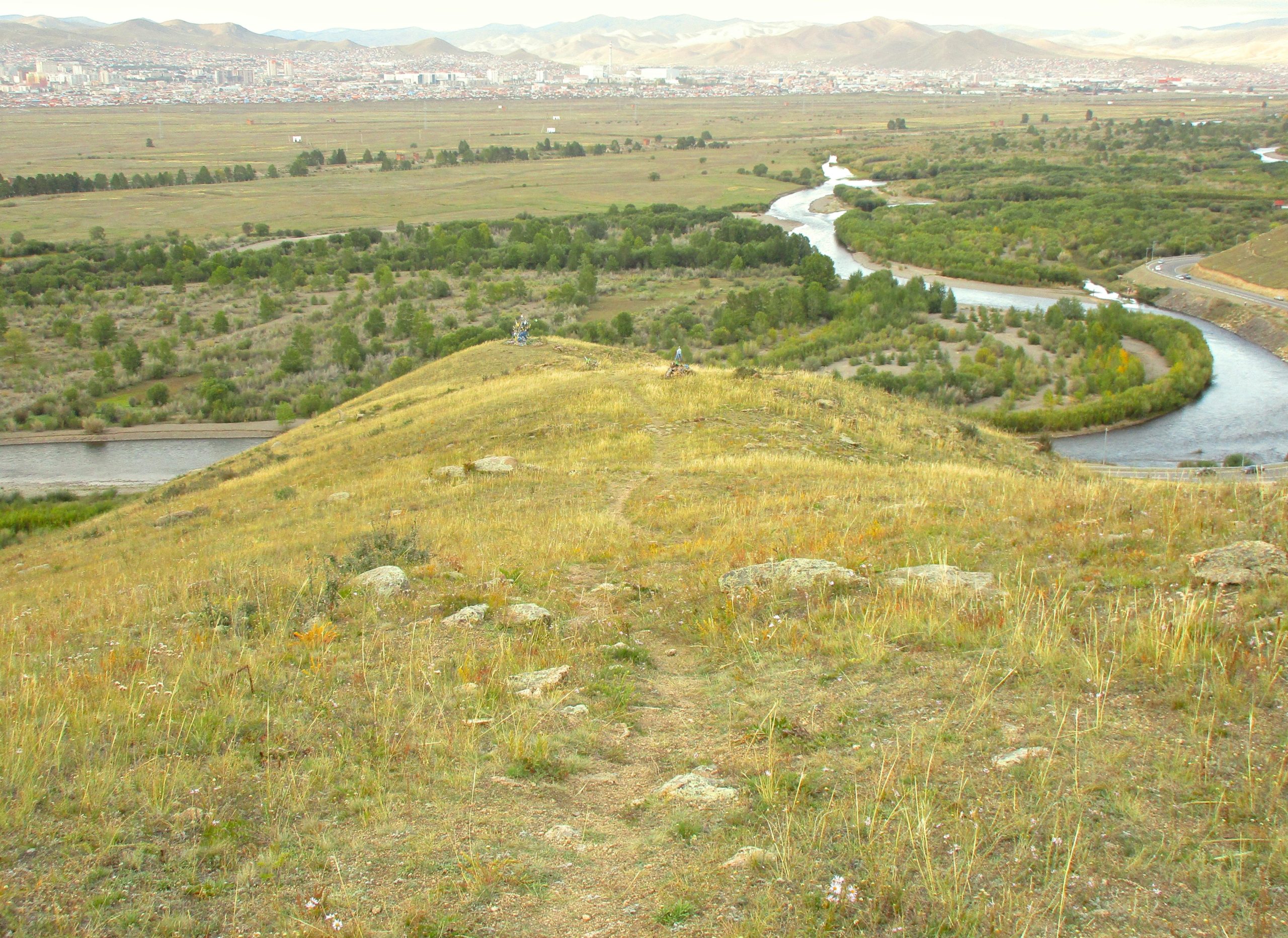 A panoramic view from a hilltop overlooking a winding river and lush greenery, with distant mountains and a cityscape in the background. The foreground features a grassy slope dotted with rocks and a dirt path leading down. The Beast mountain bike trail.