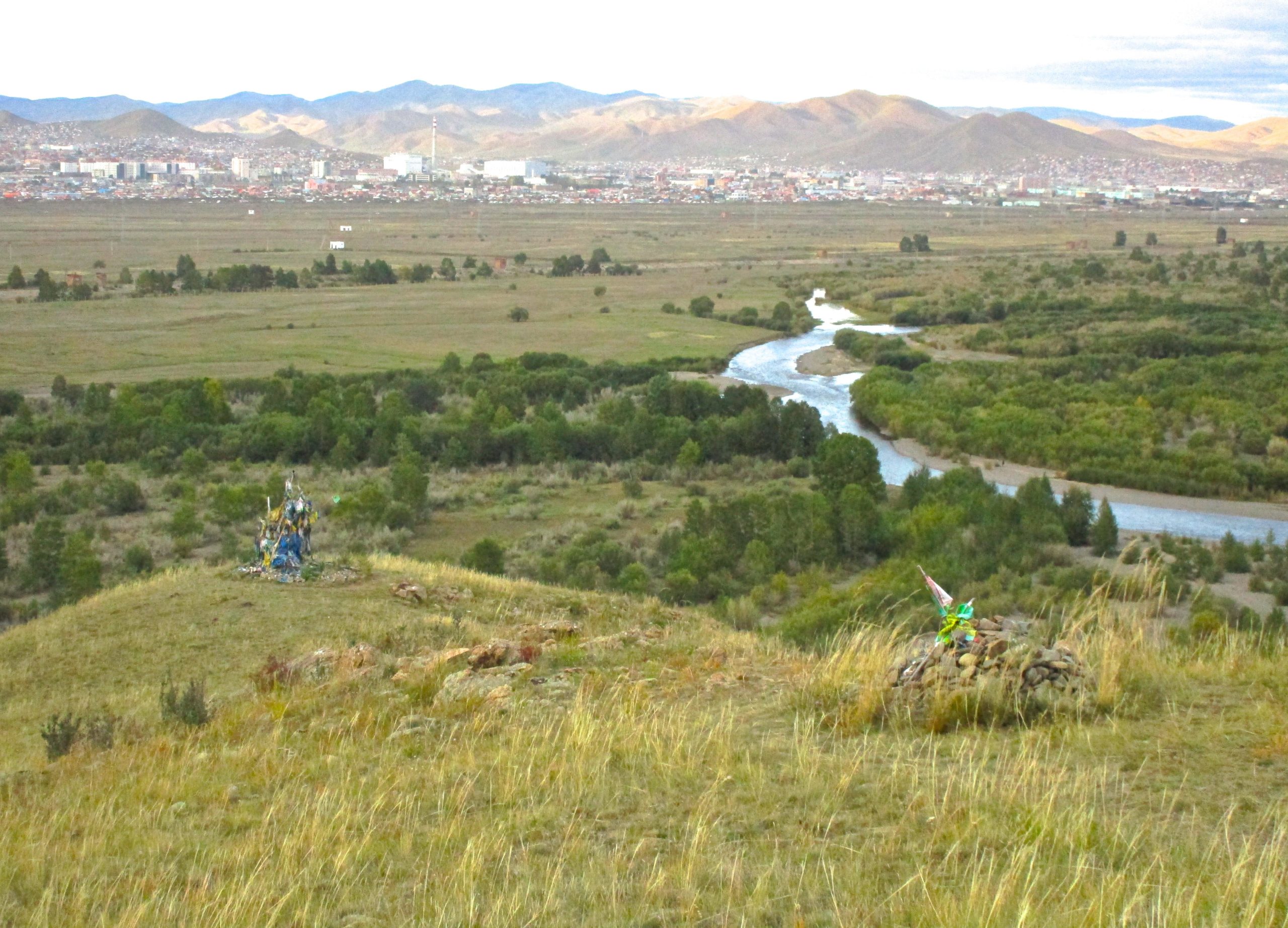 A panoramic view of a lush valley with a winding river, framed by distant mountains. In the foreground, two colorful shrines or offerings made of fabric and stones are positioned on a grassy hilltop, while a small town can be seen in the background, nestled among the rolling hills and greenery. The sky is slightly overcast, adding a serene atmosphere to the landscape. The Beast mountain bike trail.