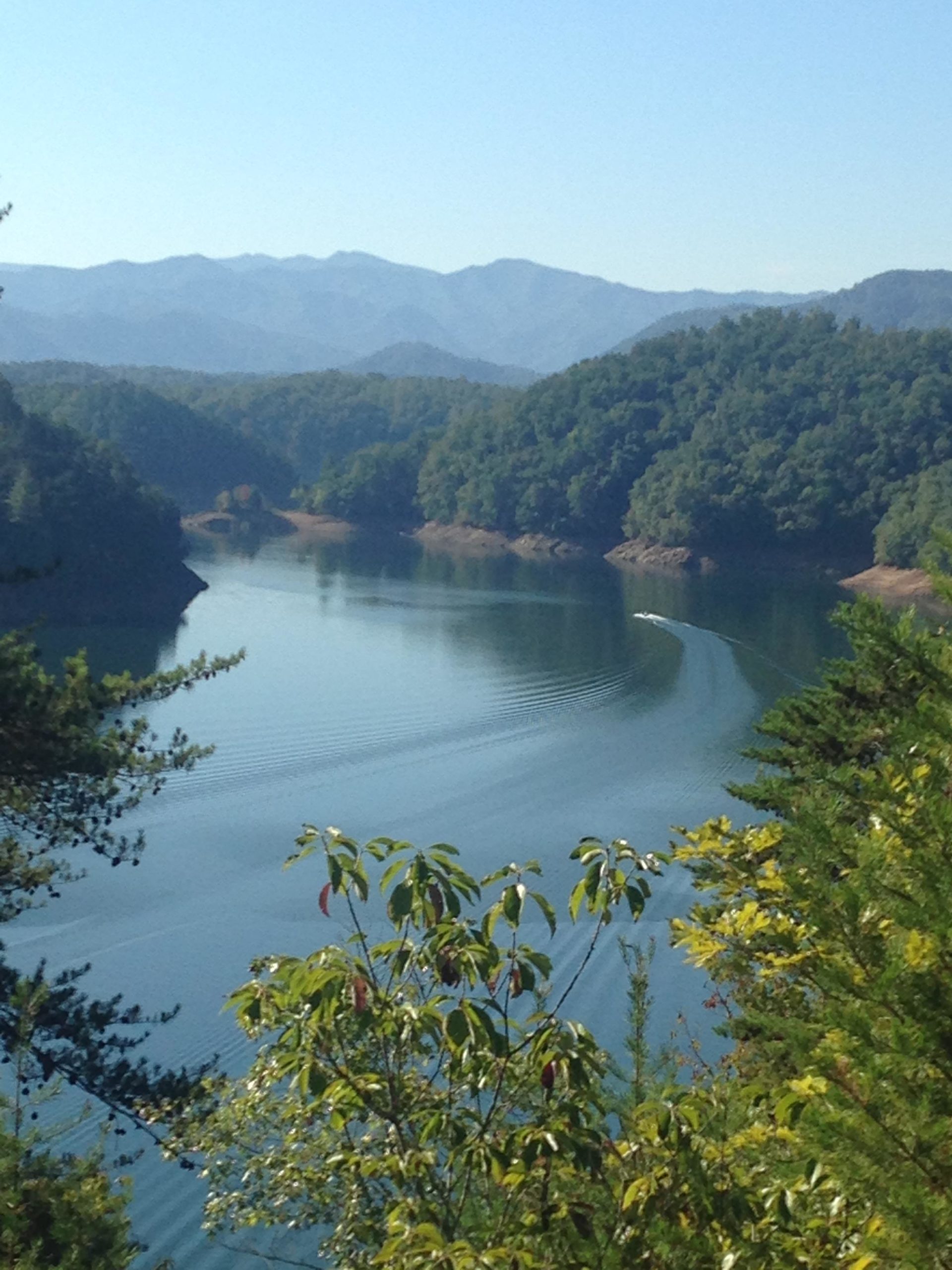 A serene view of a calm lake surrounded by lush green hills and distant mountains under a clear blue sky, with light ripples on the water's surface and colorful foliage in the foreground. Tsali Right Loop mountain bike trail.