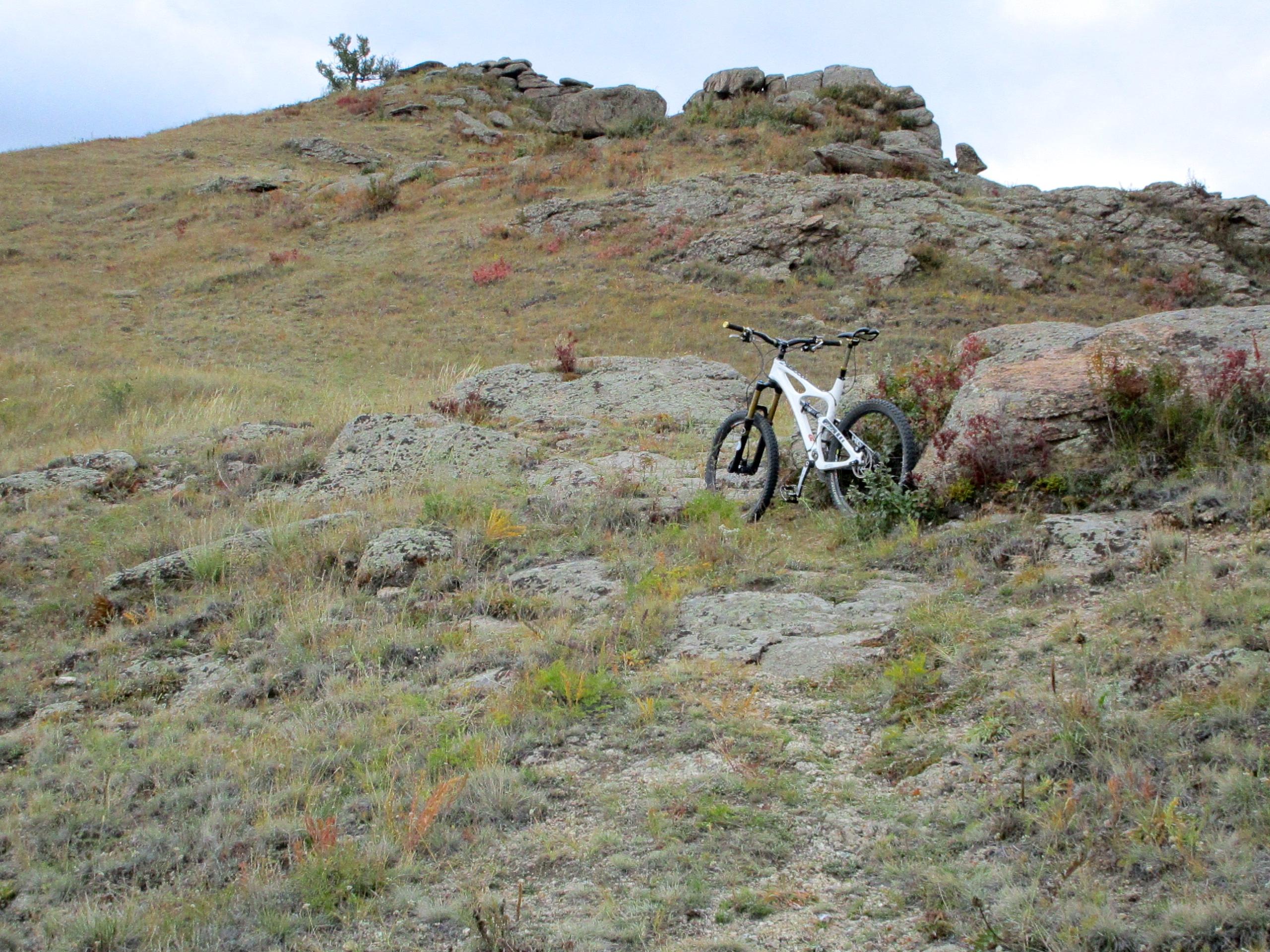 A white mountain bike is parked on rocky terrain with patches of grass and sparse vegetation under a cloudy sky. Rolling hills and boulders are visible in the background, creating a rugged outdoor scene. The Beast mountain bike trail.