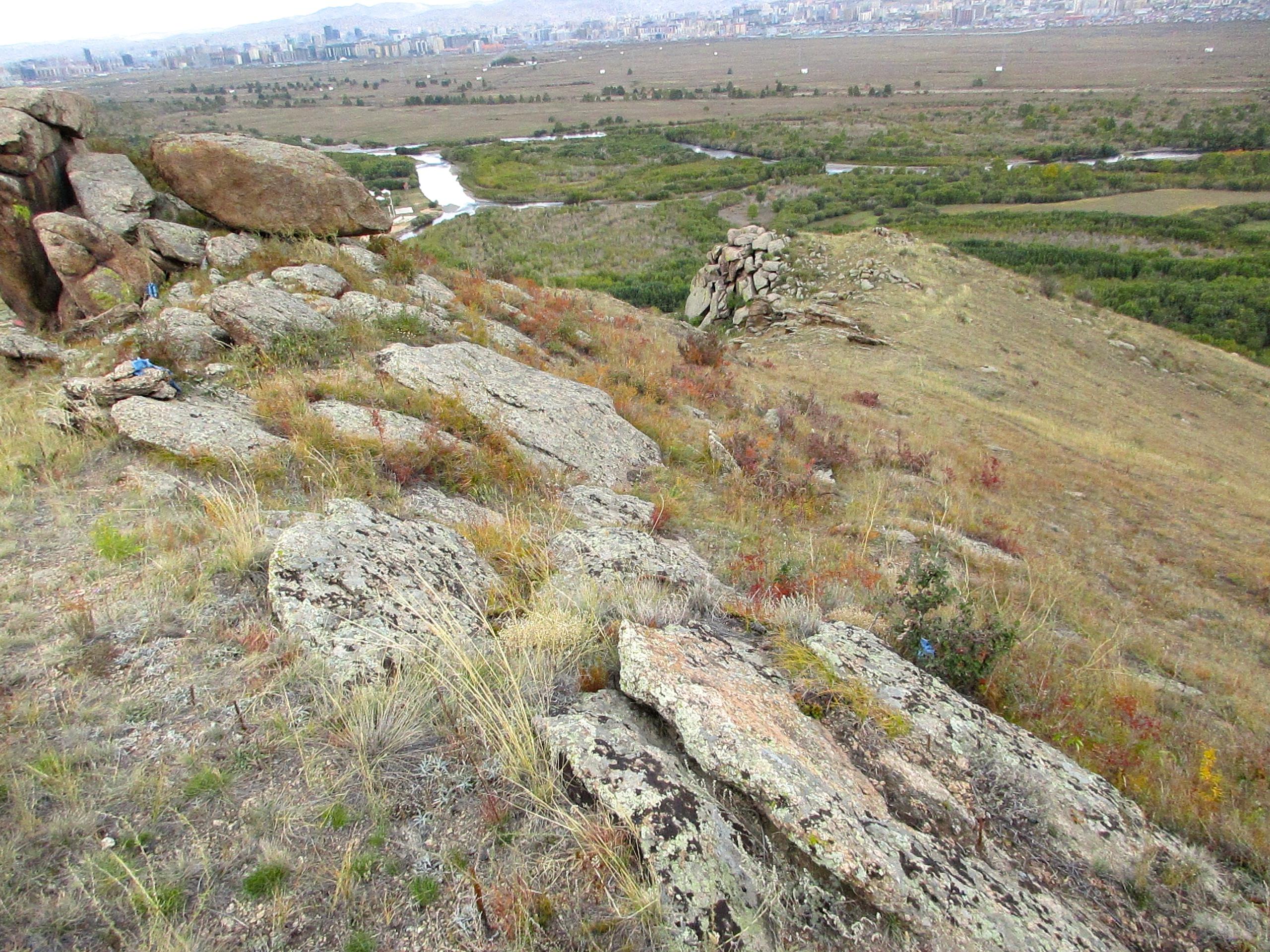A rocky hillside with scattered boulders and grass, overlooking a valley with a winding river. In the distance, a city skyline is visible against a backdrop of rolling hills and a cloudy sky. Wildflowers and shrubs add splashes of color to the landscape. The Beast mountain bike trail.