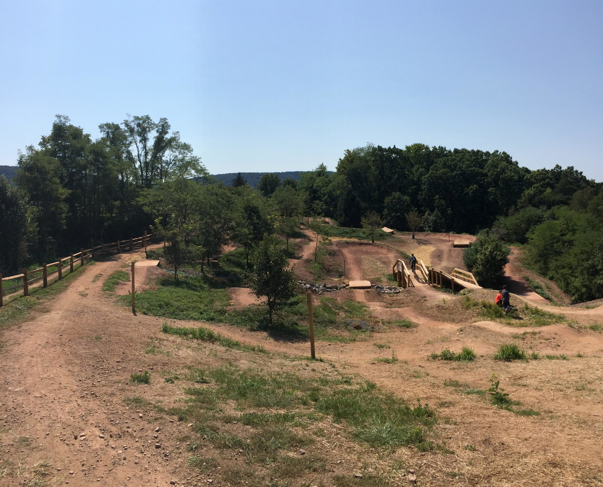 A scenic view of a dirt bike park featuring winding trails and small hills. A wooden bridge spans over a path, surrounded by lush greenery and trees. Two people can be seen enjoying the area, with a clear blue sky overhead. The landscape showcases a mix of dirt paths and grassy areas, ideal for outdoor activities. Allegrippis Trails mountain bike trail.