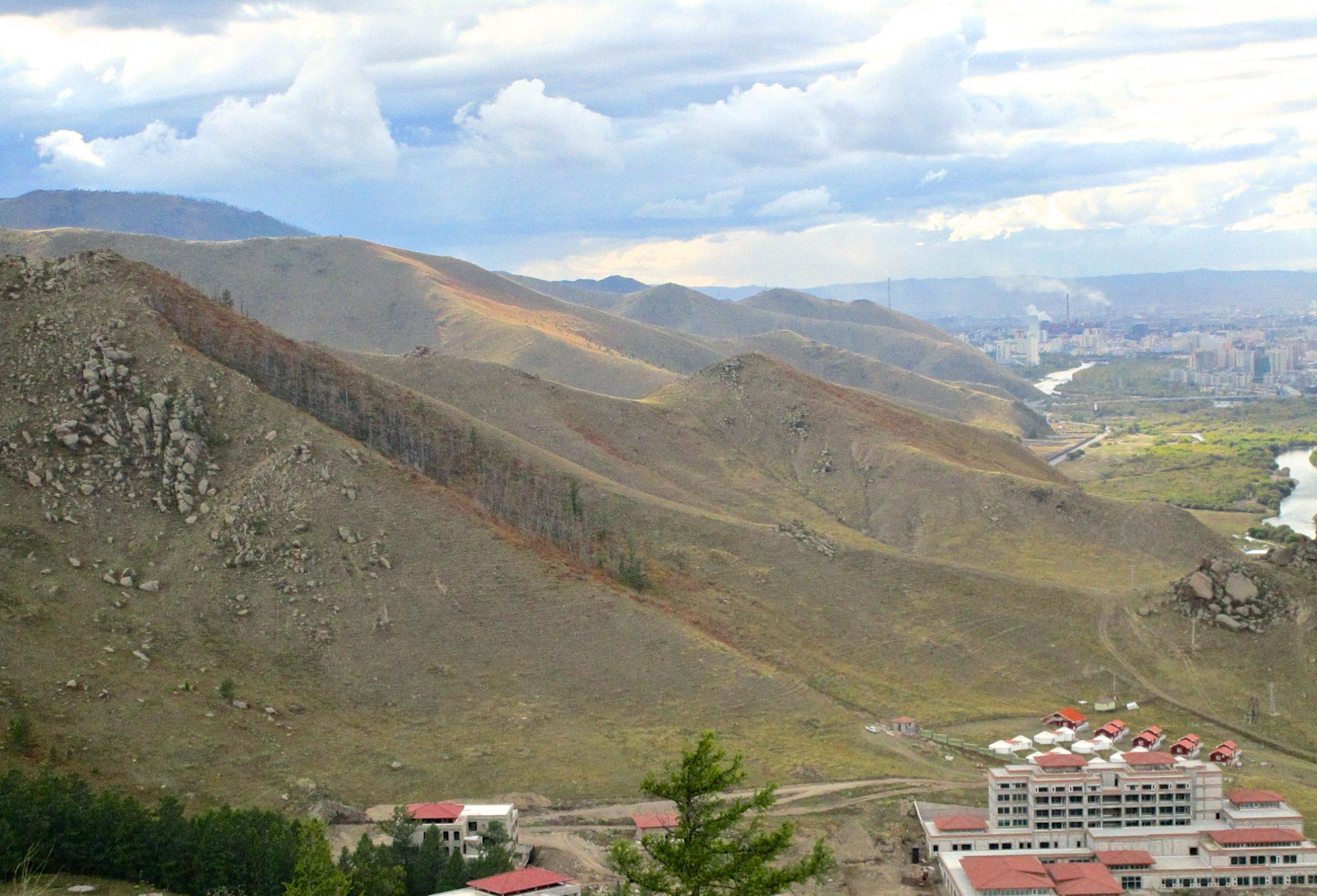 A scenic view of rolling mountains under a cloudy sky, featuring rocky slopes and sparse vegetation. In the foreground, there are buildings with red roofs, and a winding river can be seen in the distance, alongside a city skyline. The Beast mountain bike trail.