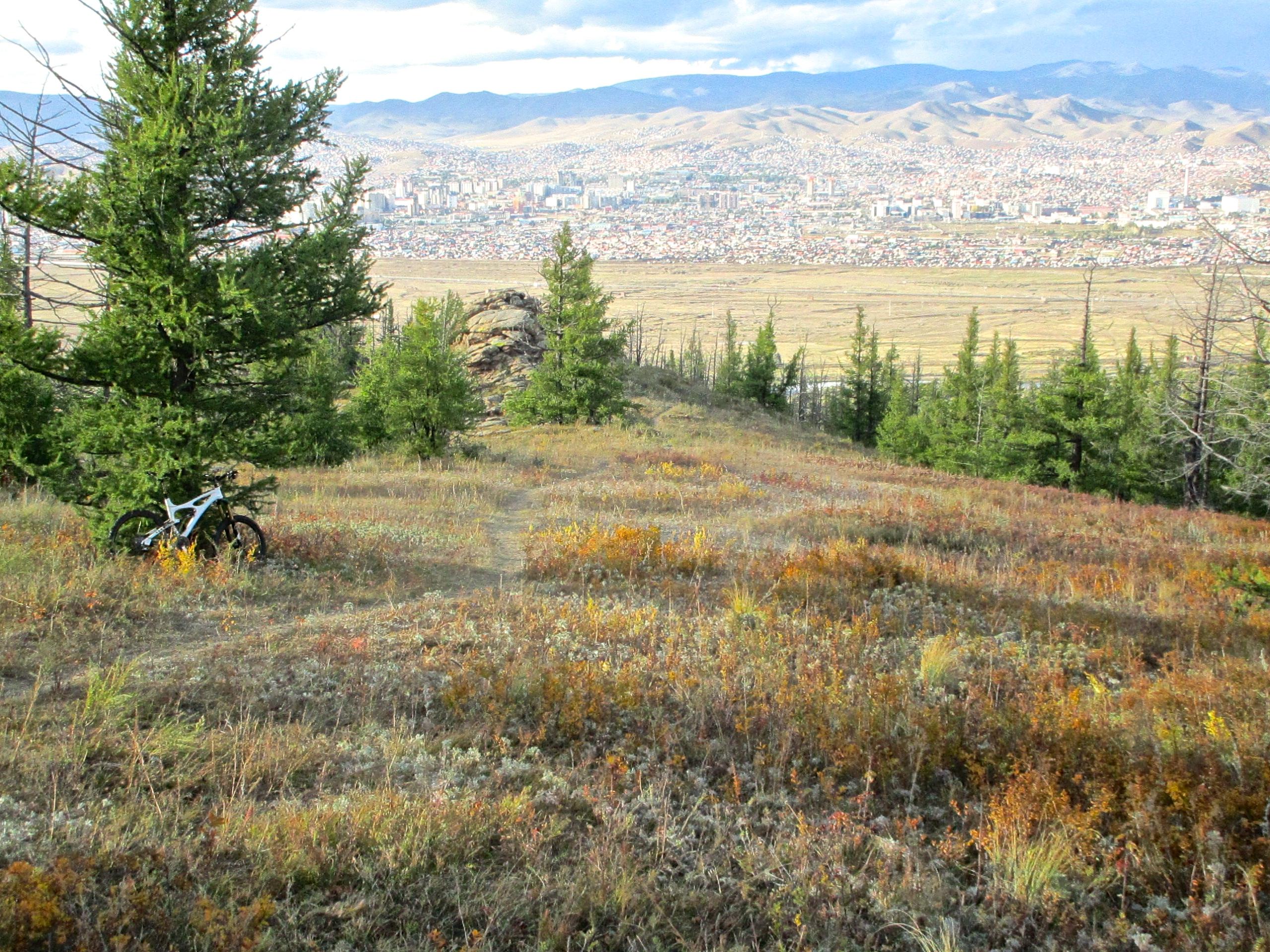 A scenic view from a hillside overlooking a city, with green pine trees and colorful autumn foliage in the foreground. A bicycle is resting against a tree, and the city skyline is visible in the distance, framed by mountains under a partly cloudy sky. The Beast mountain bike trail.