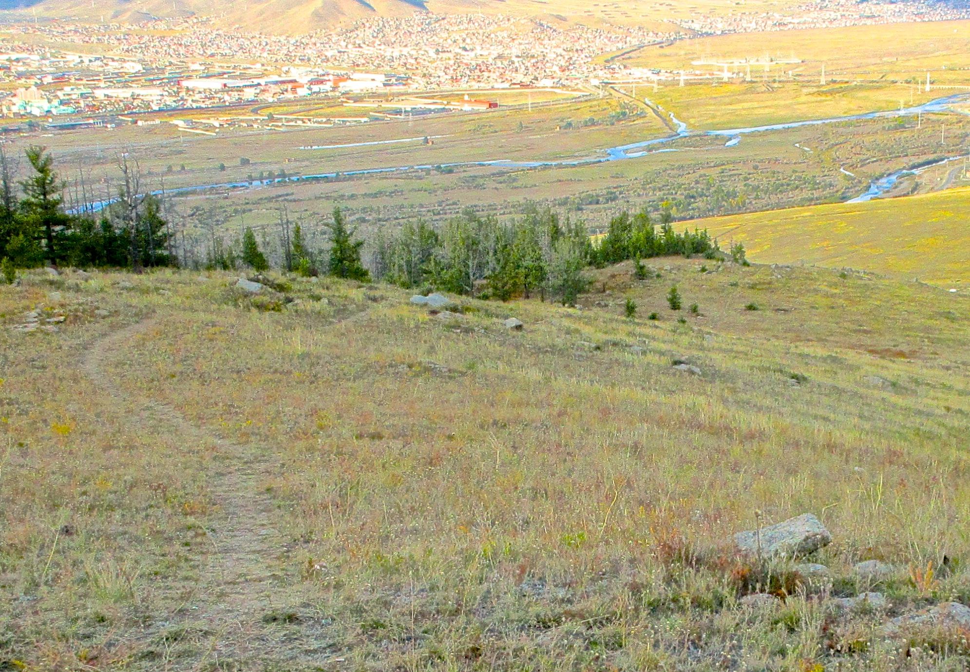 A panoramic view of a landscape featuring a grassy hillside with scattered rocks and a winding trail leading downwards. In the background, a river meanders through a valley, with a city visible on the horizon, surrounded by distant mountains. The scene captures a blend of natural and urban elements under a clear sky. The Beast mountain bike trail.