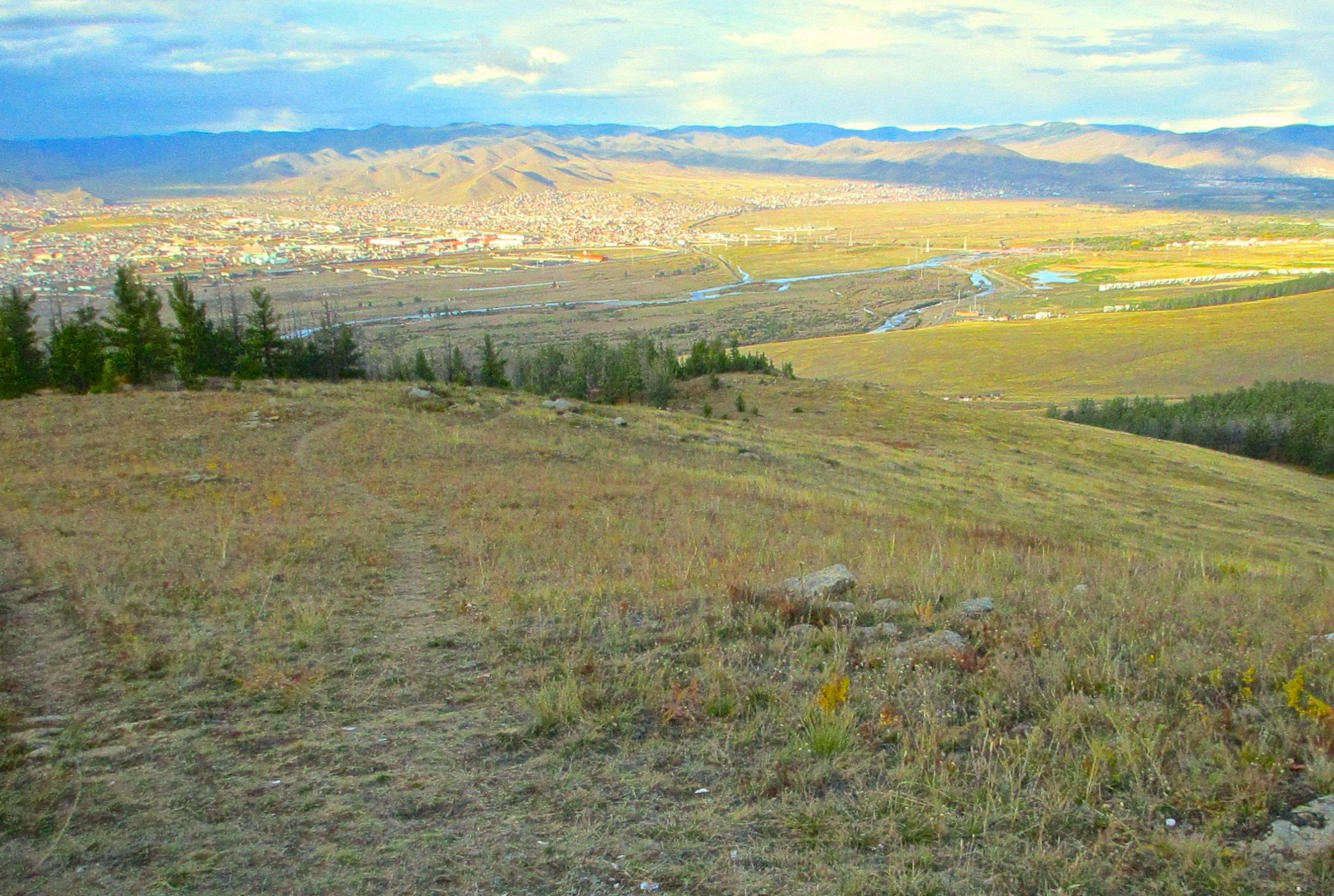 A panoramic view of a valley surrounded by mountains, showcasing a small town below with a mix of buildings and greenery. The foreground features grassy terrain with scattered rocks and trees, while the landscape offers a gentle slope leading down to the town. The sky is partly cloudy, suggesting an overcast day. The Beast mountain bike trail.