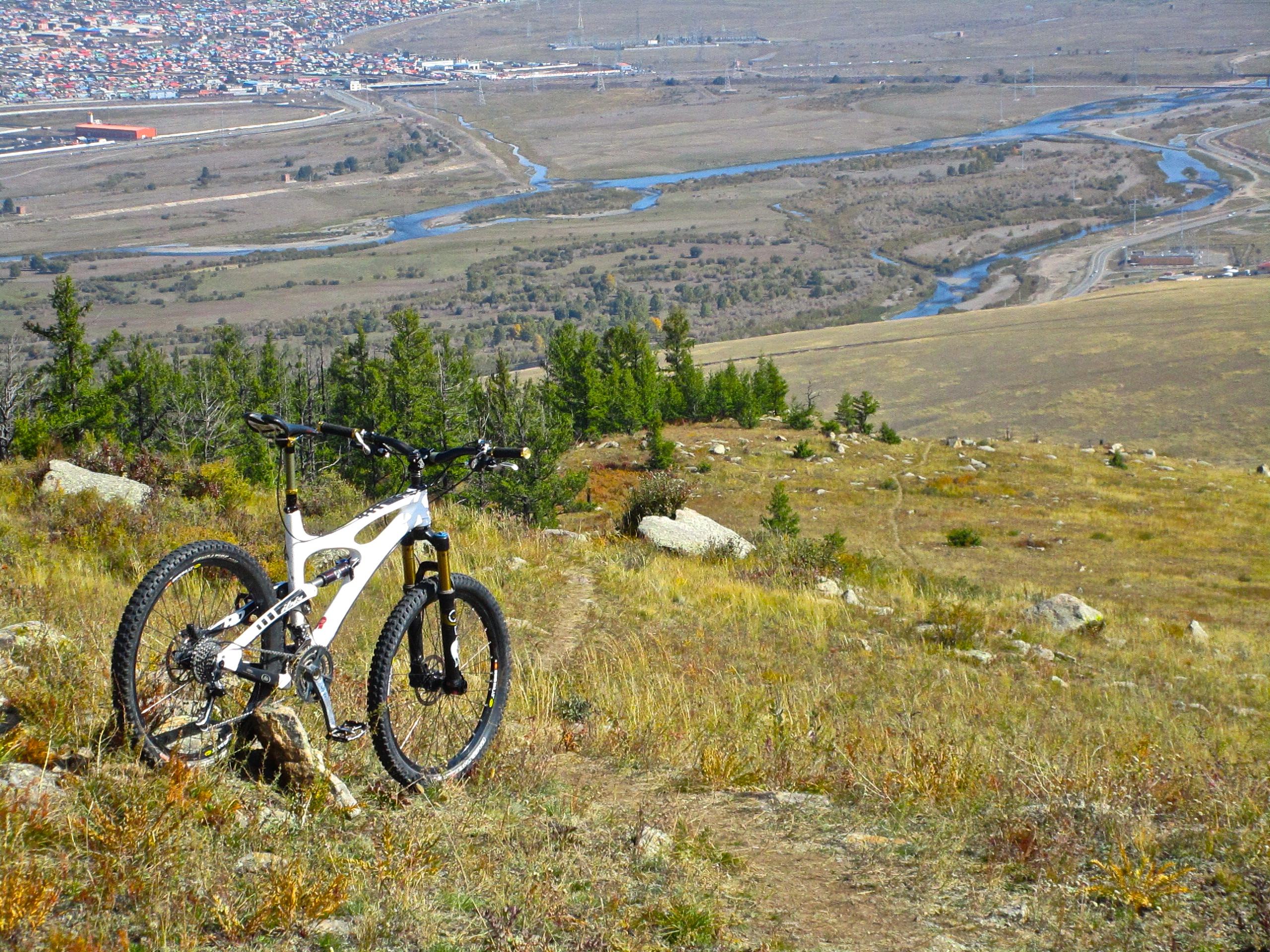 A mountain bike rests on a rocky outcrop in a grassy clearing, overlooking a winding river and a distant town below. The scene captures a vibrant landscape with scattered trees and hills, showcasing the beauty of outdoor adventure. The Beast mountain bike trail.