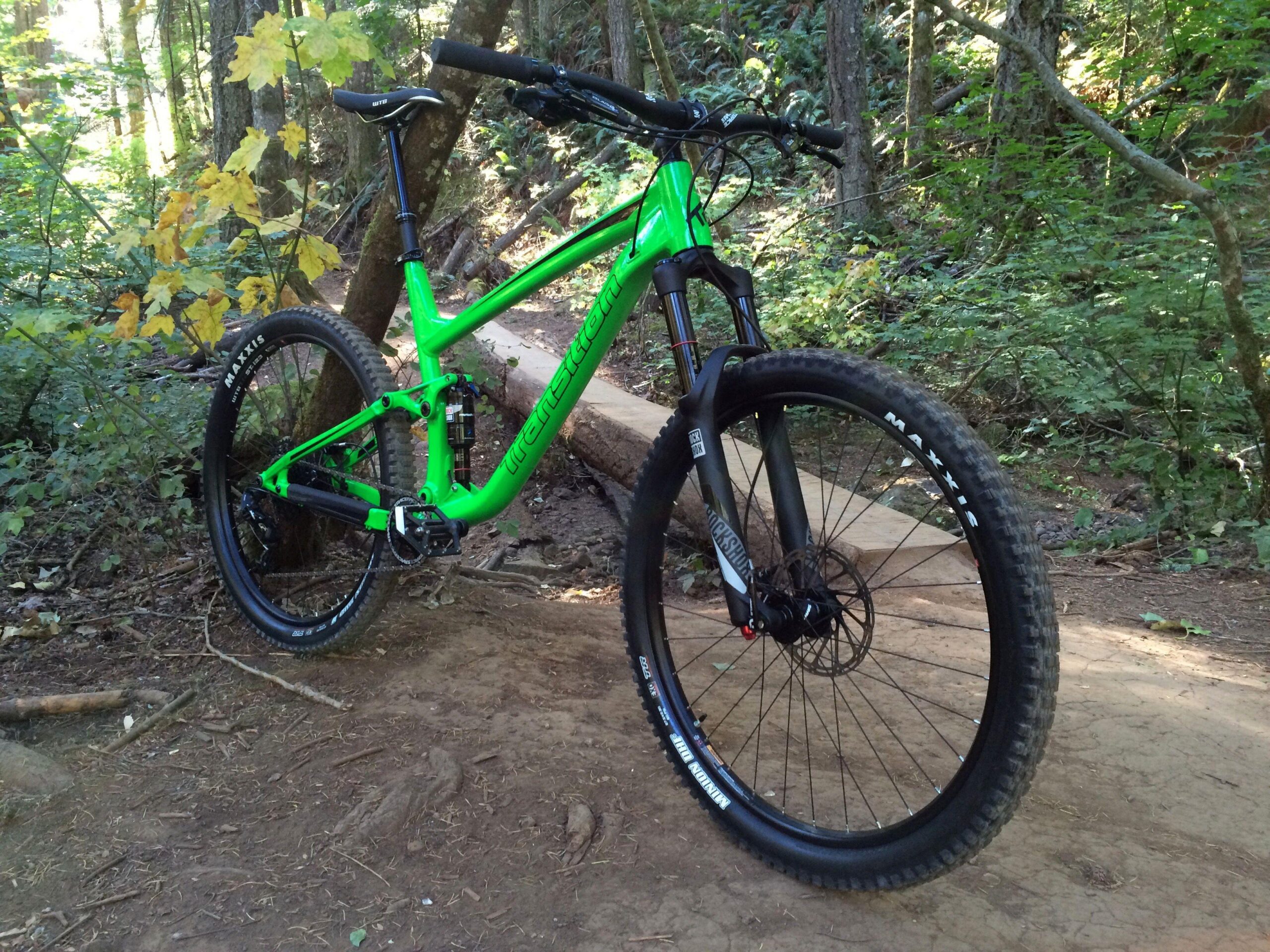 Transition Scout: A bright green mountain bike positioned on a dirt trail surrounded by lush greenery and trees. The bike features thick, knobby tires and a sturdy frame, resting next to a wooden bridge that spans the trail. Sunlight filters through the trees, creating a natural and vibrant outdoor environment.