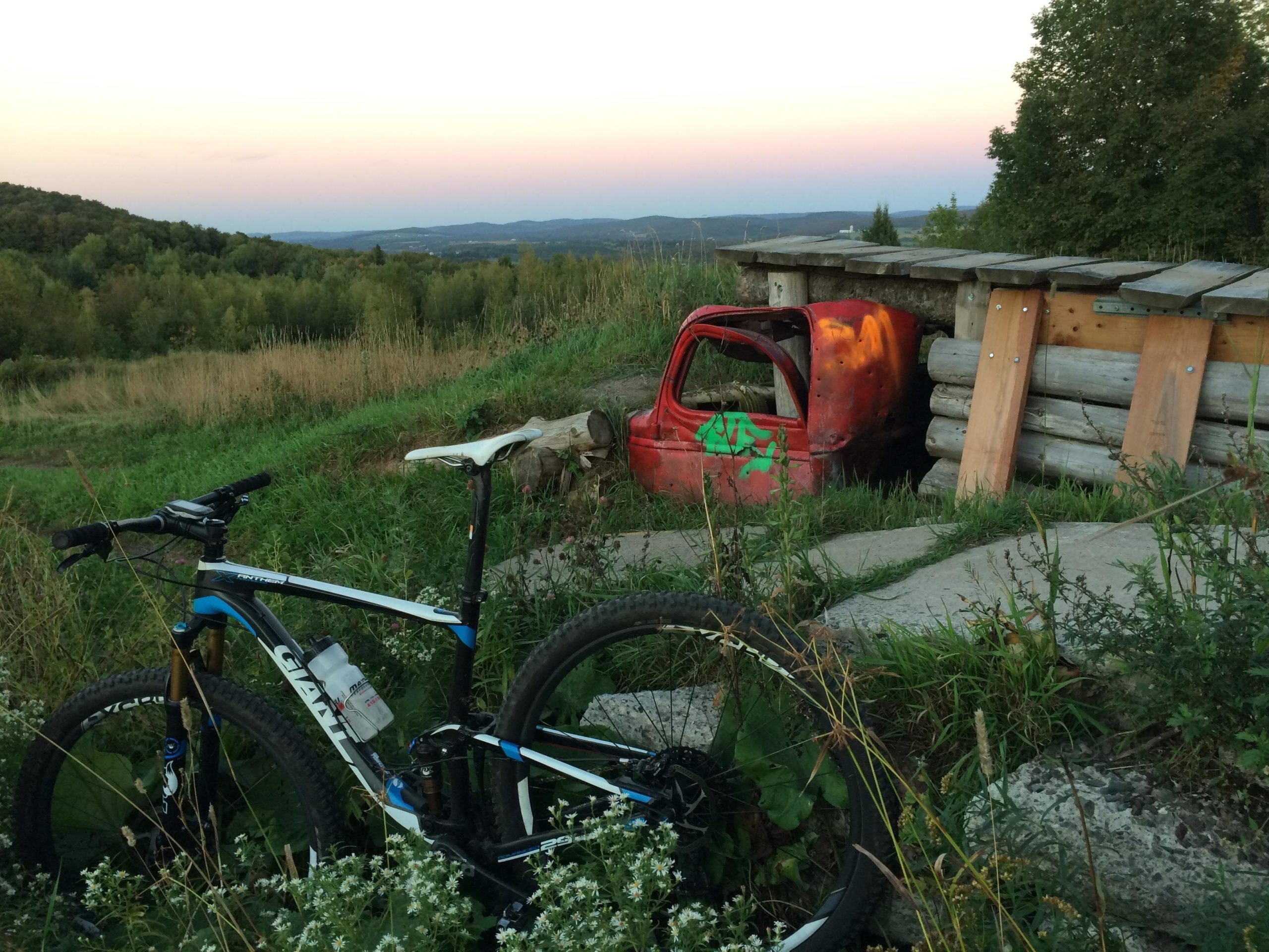 A mountain bike rests on a grassy area with wildflowers in the foreground, beside a partially visible old red truck partially obscured by a wooden structure. The backdrop features rolling hills and a sunset sky in soft pastel colors. Mont Arthabaska mountain bike trail.