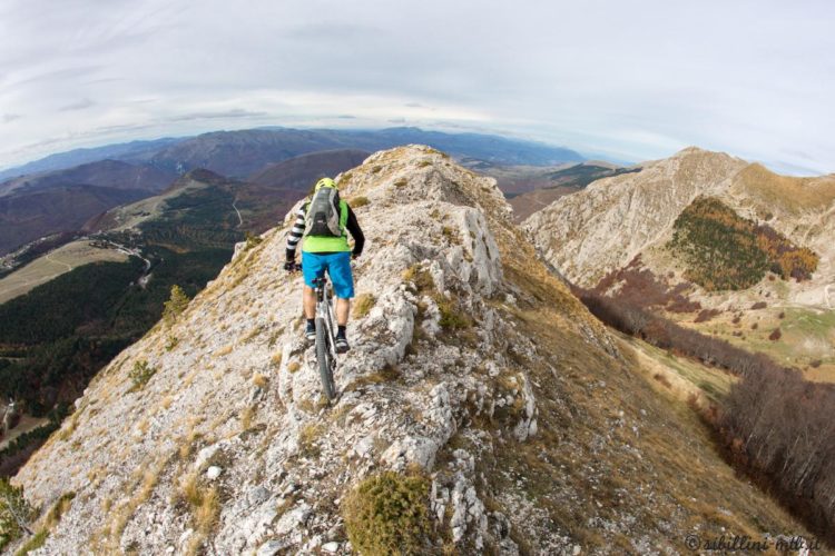 A mountain biker navigates a narrow rocky ridge with a vast valley and mountains in the background, showcasing a dramatic landscape under a cloudy sky.