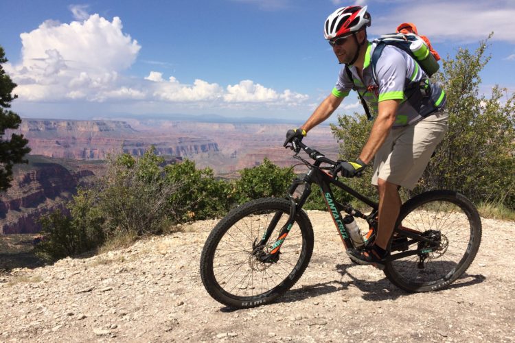 A mountain biker in a helmet and gear rides along a gravel path with a scenic view of the Grand Canyon in the background. The sky is partly cloudy, and lush green vegetation is present along the trail.