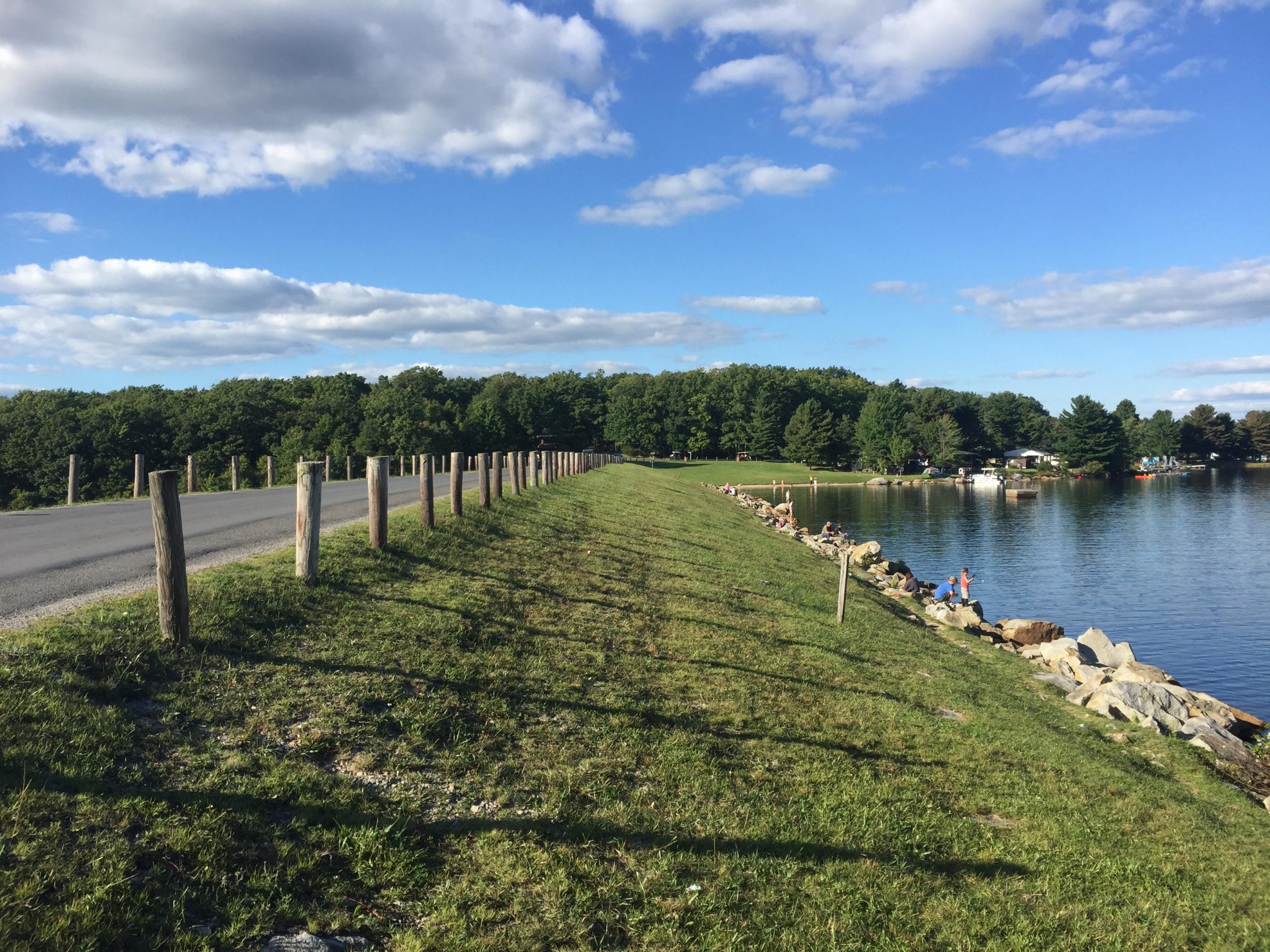A serene lakeside view featuring a grassy path lined with wooden posts, bordered by a calm body of water reflecting the blue sky and fluffy clouds. Trees provide a lush backdrop, while people can be seen fishing or enjoying the scenery along the rocky shoreline. Big Bear Lake Trail Center mountain bike trail.