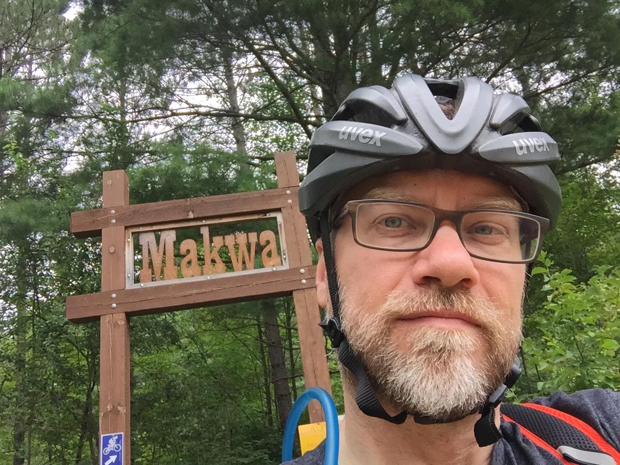 A person wearing glasses and a black bicycle helmet stands in front of a wooden sign that reads "Makwa," surrounded by greenery. Makwa Trail mountain bike trail.