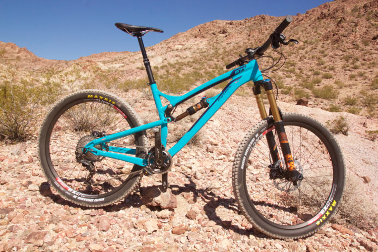 A turquoise mountain bike positioned on rocky terrain, with a clear blue sky in the background. The bike features a full suspension system, knobby tires for traction, and is set against earthy, desert-like surroundings.