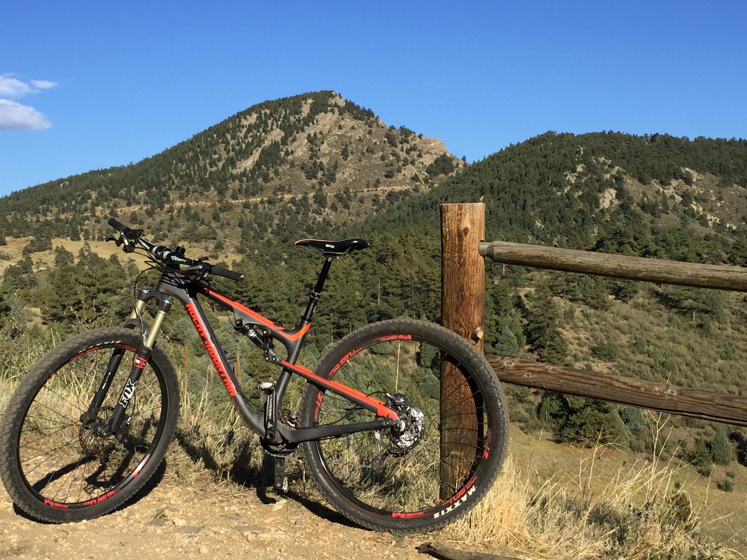 A mountain bike rests beside a wooden fence on a trail, with a scenic view of a mountainous landscape in the background. The bike has a black and orange frame, and the terrain is covered with greenery and trees under a clear blue sky. Centennial Cone Park mountain bike trail.