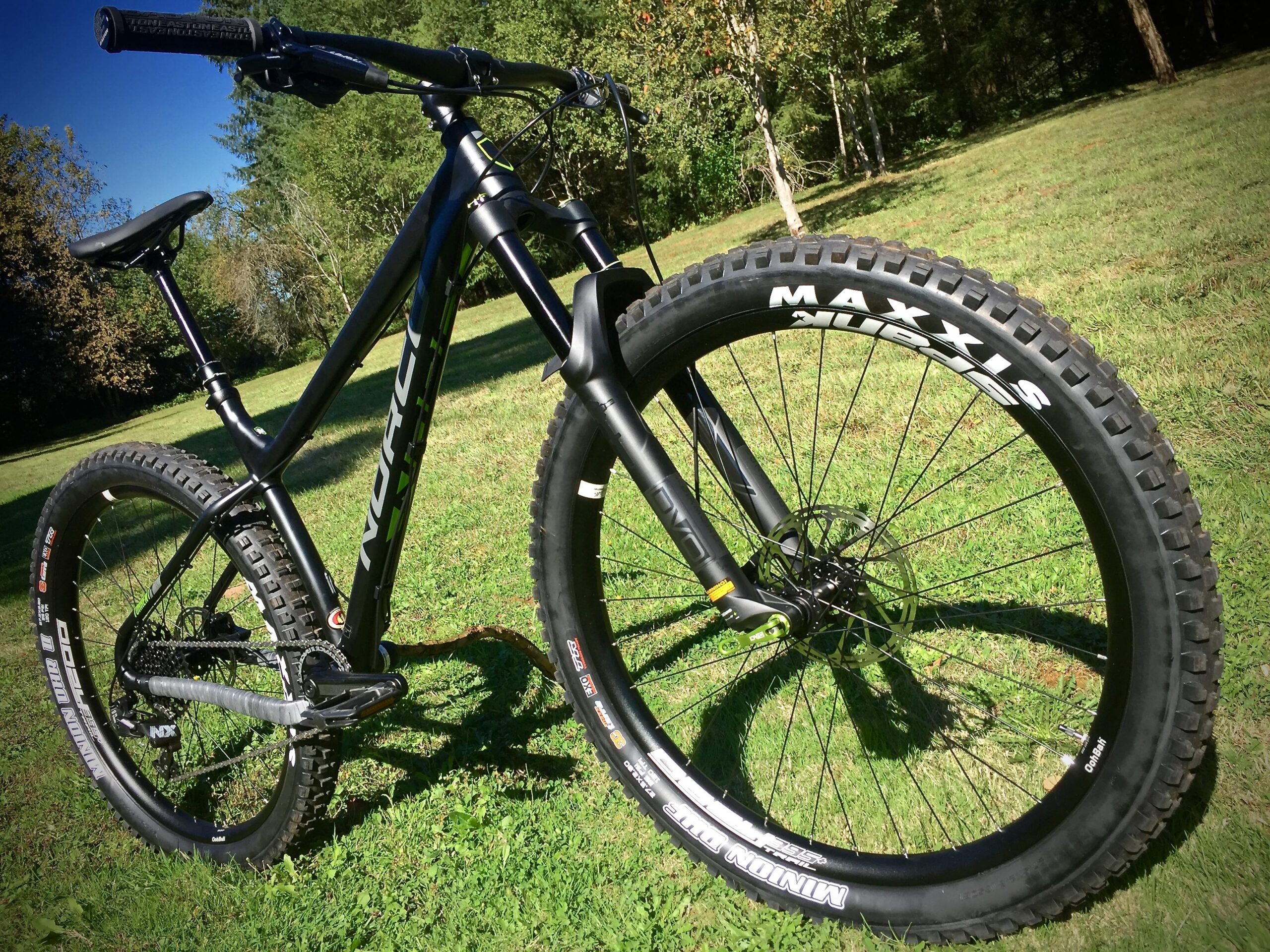 Norco Torrent 7: A close-up view of a black mountain bike with green accents, featuring large Maxxis tires and a sturdy frame. The bike is positioned on a grassy area surrounded by trees, showcasing its design and rugged features.