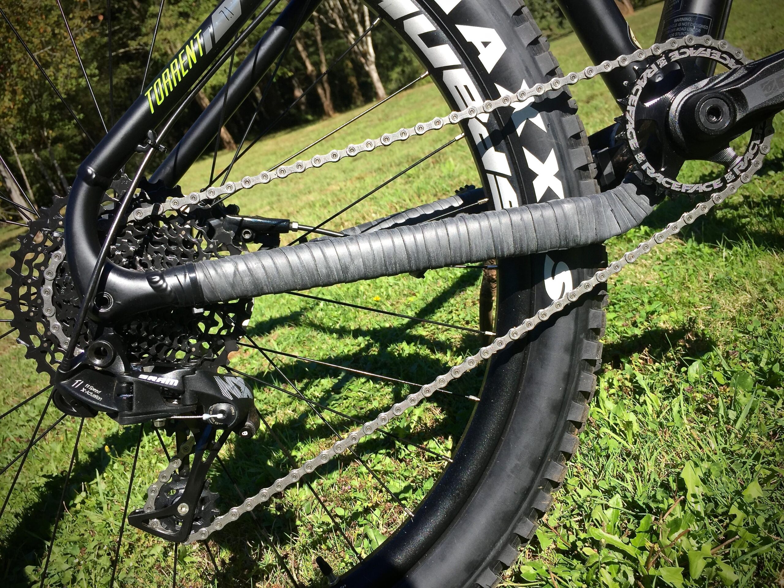 Norco Torrent 7: Close-up view of a mountain bike's drivetrain, showcasing the cassette, derailleur, and chain, with the rear wheel and tire visible in the background. The image is set against a grassy landscape.
