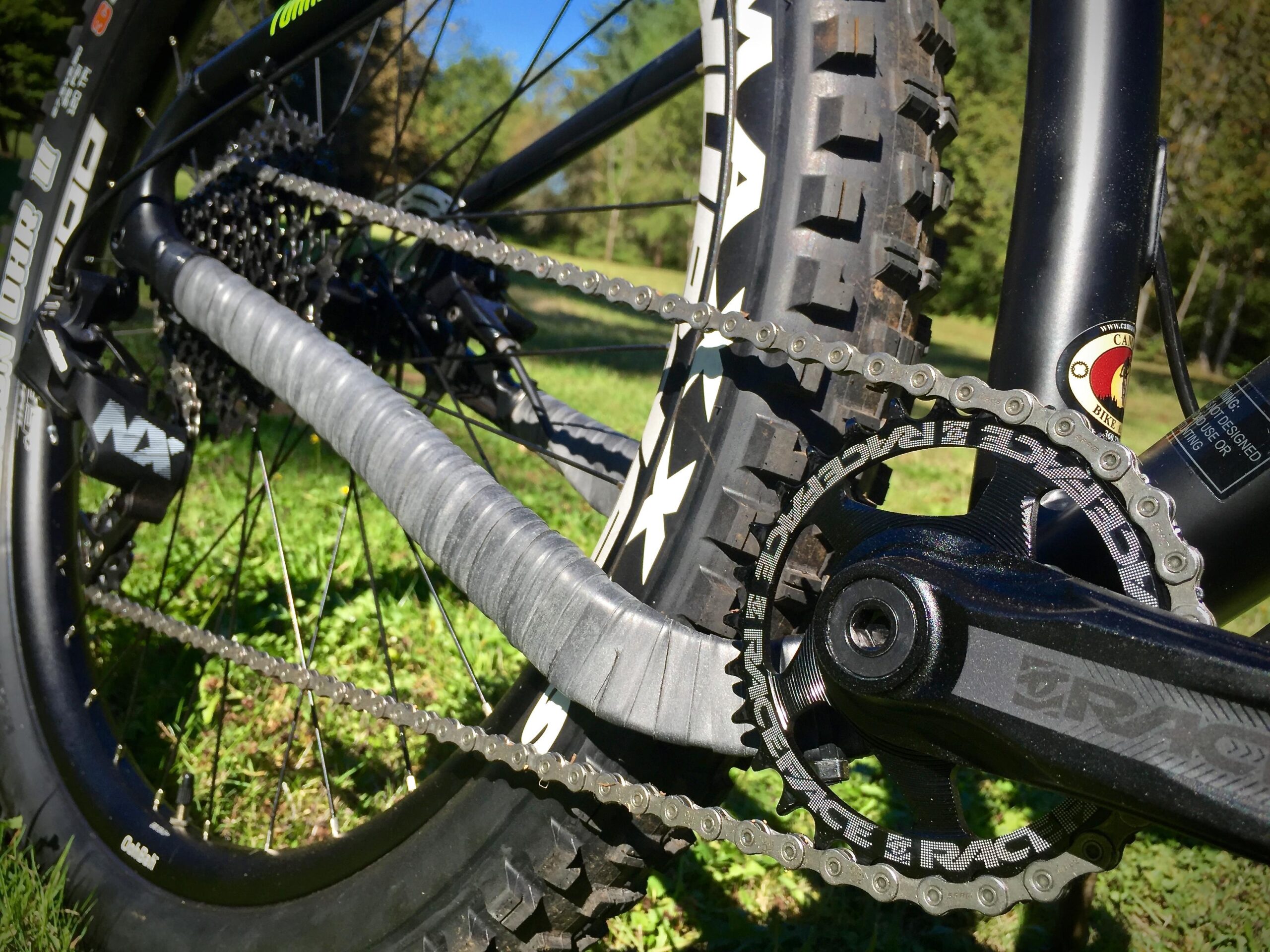 Norco Torrent 7: Close-up view of a mountain bike's drivetrain, featuring a chain, rear cassette, and crankset, with a black tire in the background. The scene is set against a green grassy area under a clear blue sky.