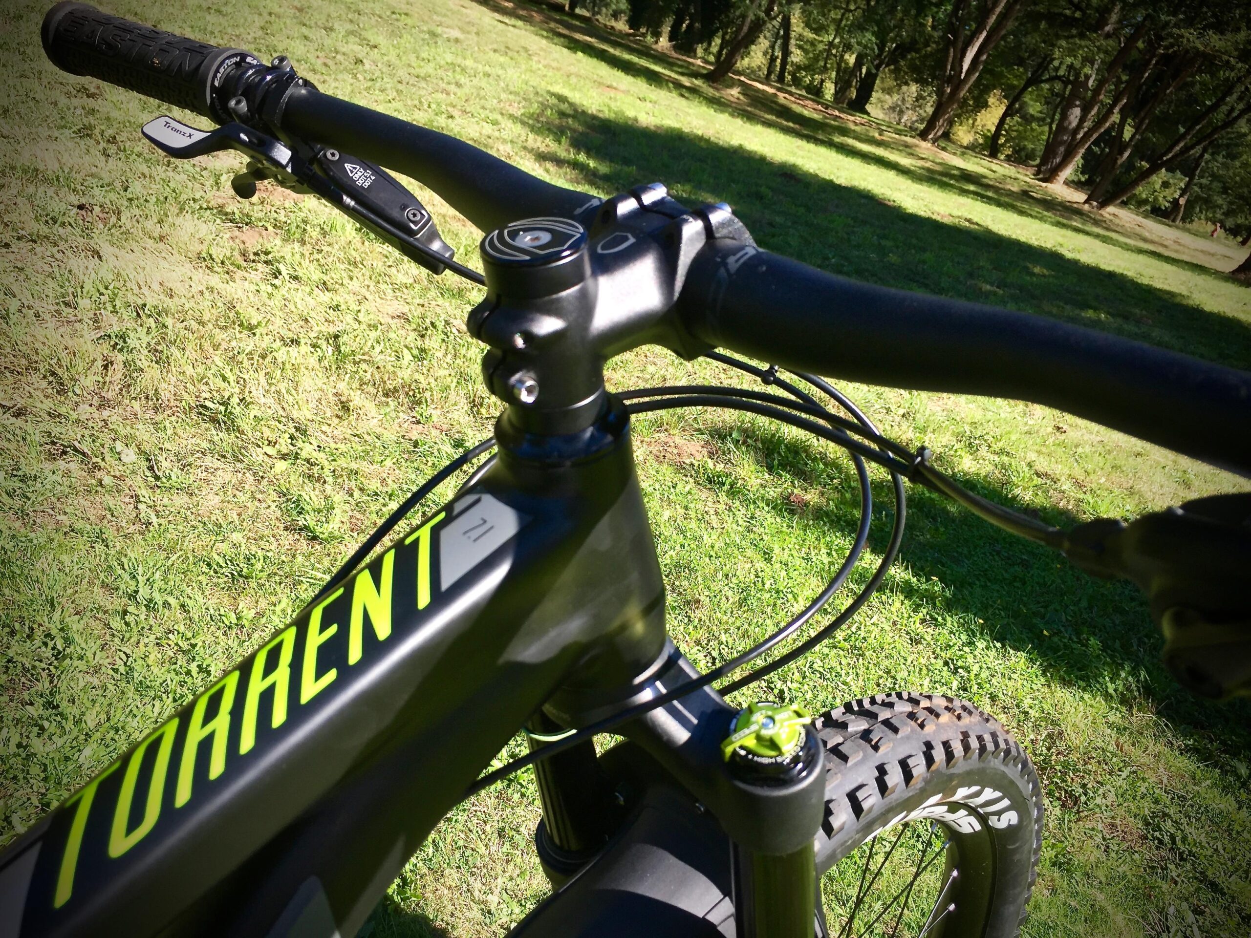 Norco Torrent 7: Close-up view of a mountain bike's handlebars and frame, featuring the word "TORRENT" in bright green. The scene is set in a grassy area with trees in the background, highlighting the bike's components, including the brake lever and gear shifters.