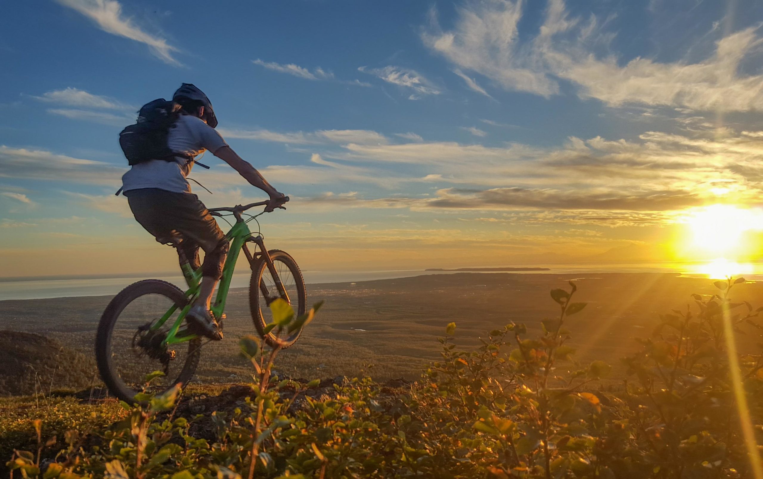 A mountain biker rides along a rocky trail at sunset, with a panoramic view of valleys and a body of water in the background. The sky is painted with warm colors, while bushes and foliage frame the scene in the foreground. The Dome/ Knoya Peak mountain bike trail.