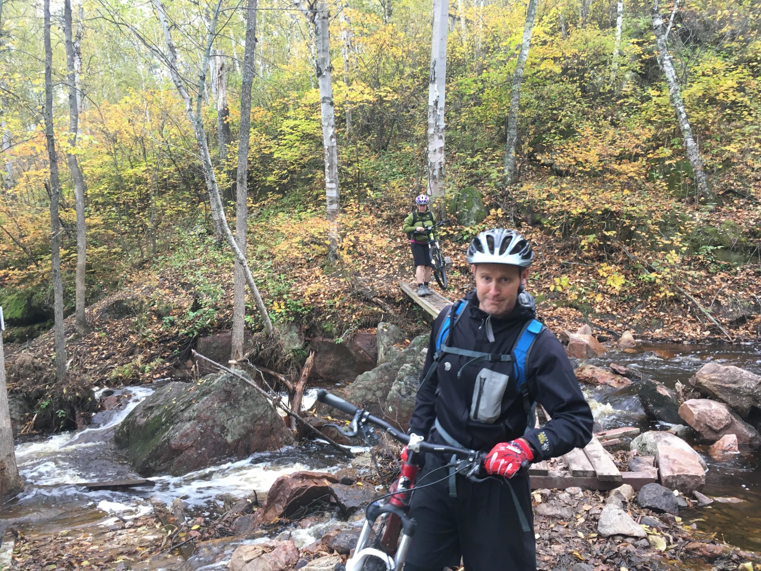 Two mountain bikers pause along a wooded path beside a flowing stream. One rider, dressed in a black outfit with a helmet, stands in the foreground holding a bicycle. The second rider is positioned on a wooden bridge in the background. The scene is set in a colorful autumn landscape with vibrant yellow and green foliage. The Blue Highway mountain bike trail.