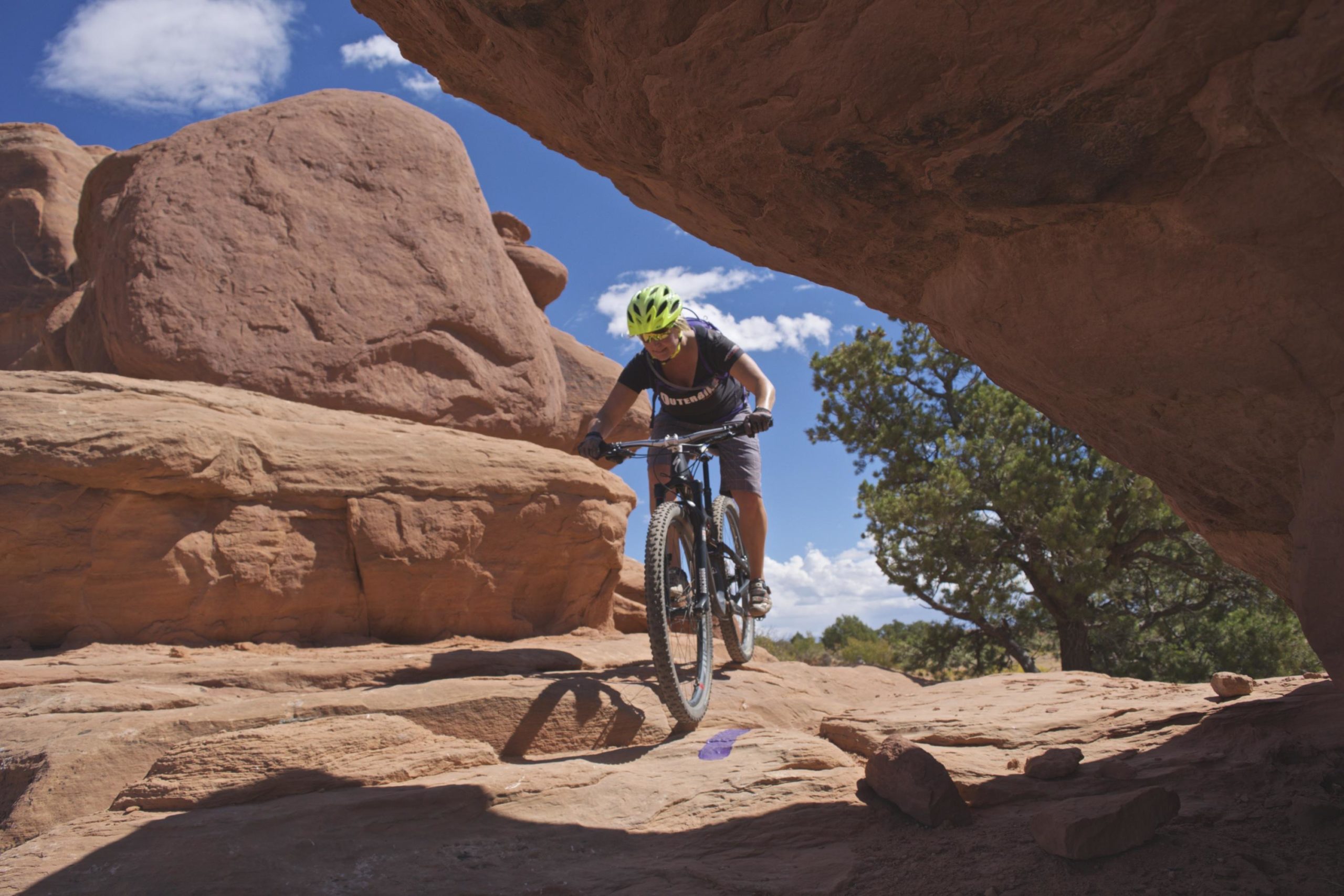 A mountain biker navigating rocky terrain under a blue sky with clouds, surrounded by large rock formations and greenery in a natural landscape. Navajo Rocks mountain bike trail.
