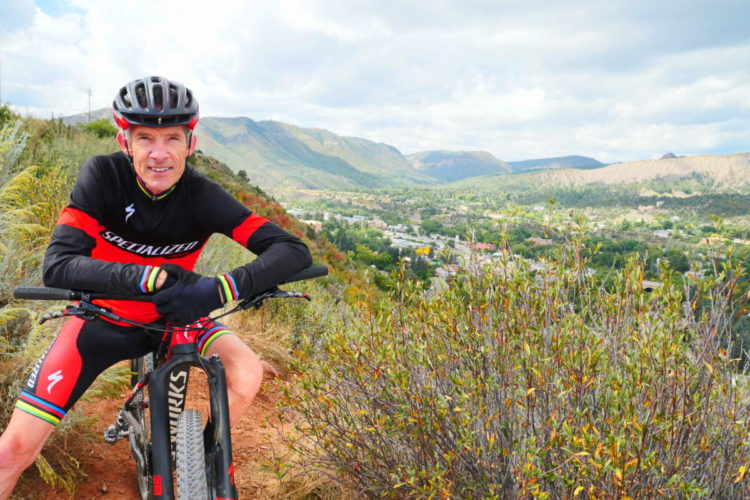 A mountain biker in a red and black Specialized cycling outfit poses with his bike on a scenic trail overlooking a lush green valley and mountains in the background. The sky is partly cloudy, creating a vibrant outdoor atmosphere.
