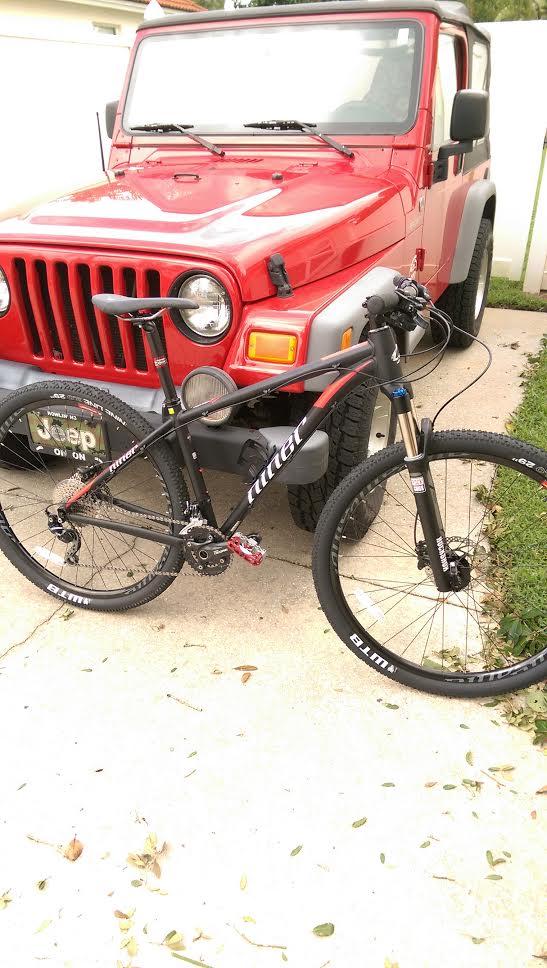 Niner EMD 9: Alt text: A black mountain bike leaned against a red Jeep Wrangler parked on a concrete driveway, surrounded by scattered leaves and grass.