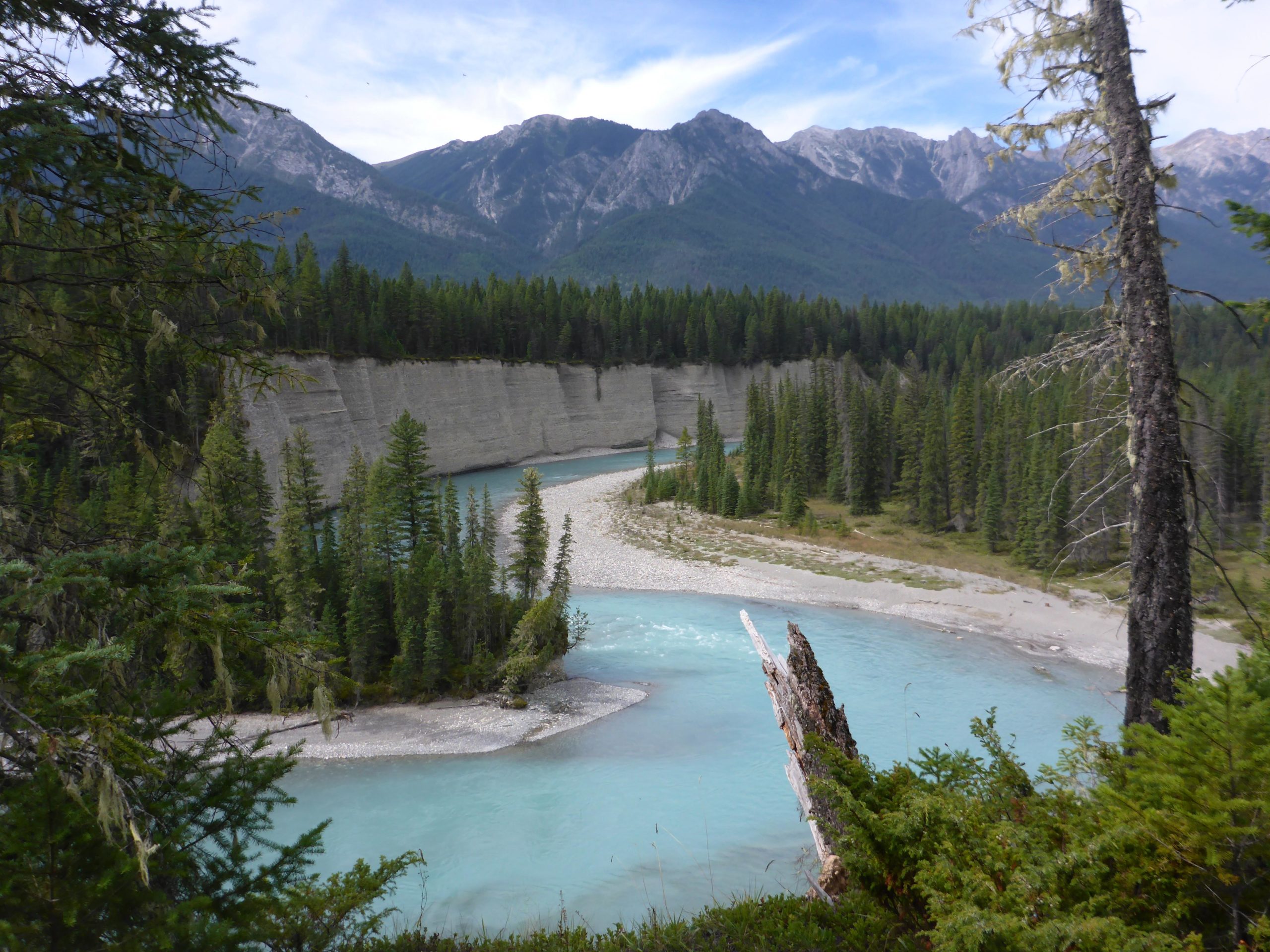 A serene landscape featuring a winding river with turquoise water surrounded by lush green forests and tall mountains in the background. The riverbanks are lined with smooth pebbles, and the scene is bathed in natural light, highlighting the beauty of the wilderness. Nipika Mountain Resort mountain bike trail.