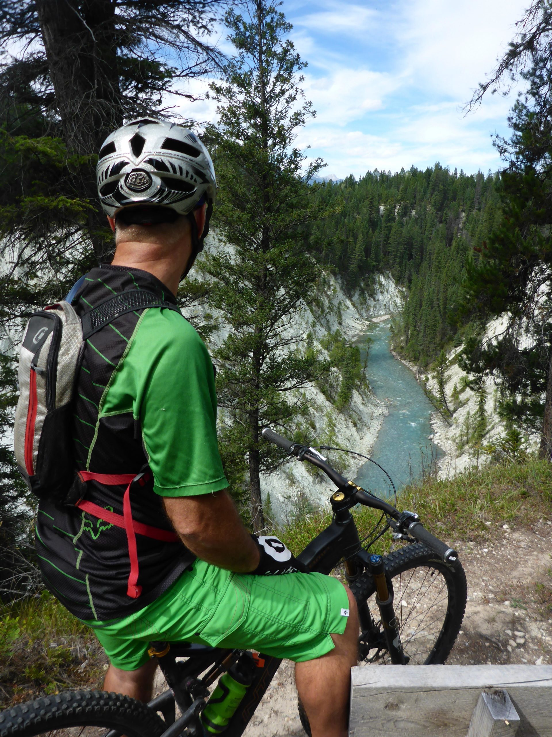 A mountain biker wearing a green jersey and shorts sits on a bike, overlooking a scenic river valley surrounded by dense evergreen trees and clear blue skies. The rider is positioned on a rocky outcrop, contemplating the trail ahead. Nipika Mountain Resort mountain bike trail.