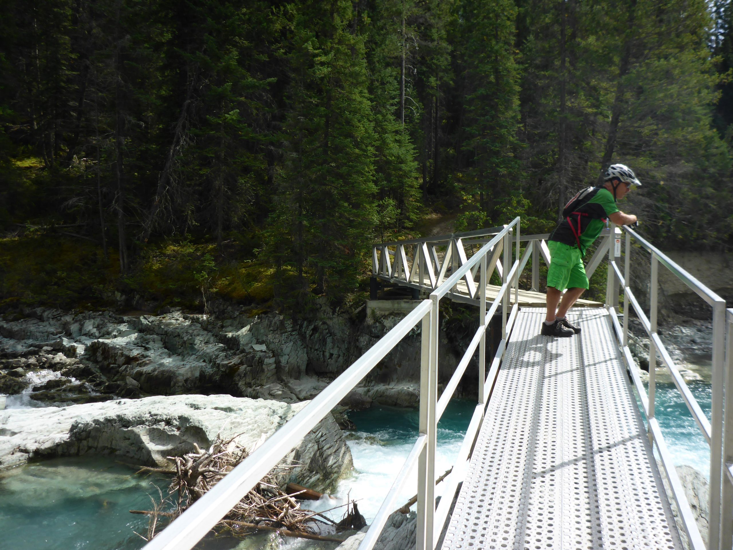 A person dressed in green shorts and a helmet leans over a metal bridge overlooking a rocky river surrounded by dense green trees. Nipika Mountain Resort mountain bike trail.