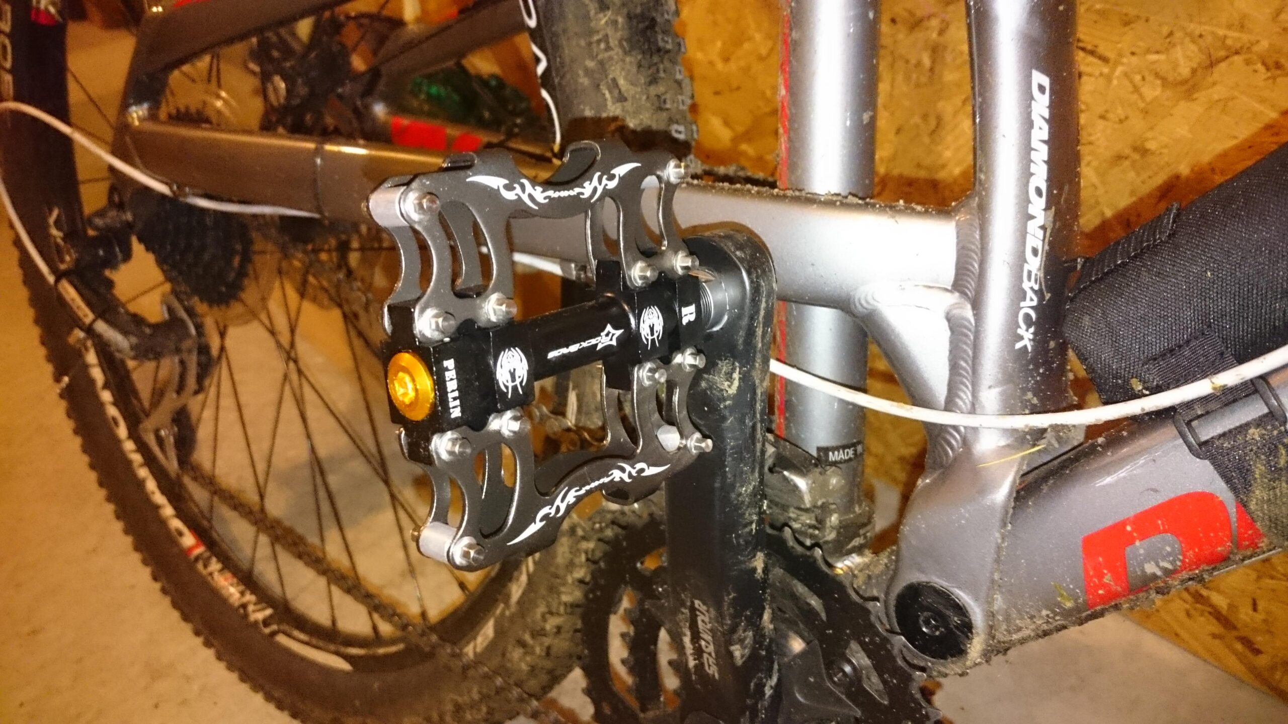 Diamondback Recoil 29: Close-up view of a mountain bike's drivetrain, featuring a detailed shot of the pedal and crankset. The pedal is black with silver accents, mounted on a gray bike frame with the brand "Diamondback" visible. The bike shows signs of use, with dirt and mud on the frame and components, indicating it's been recently ridden on rough terrain.