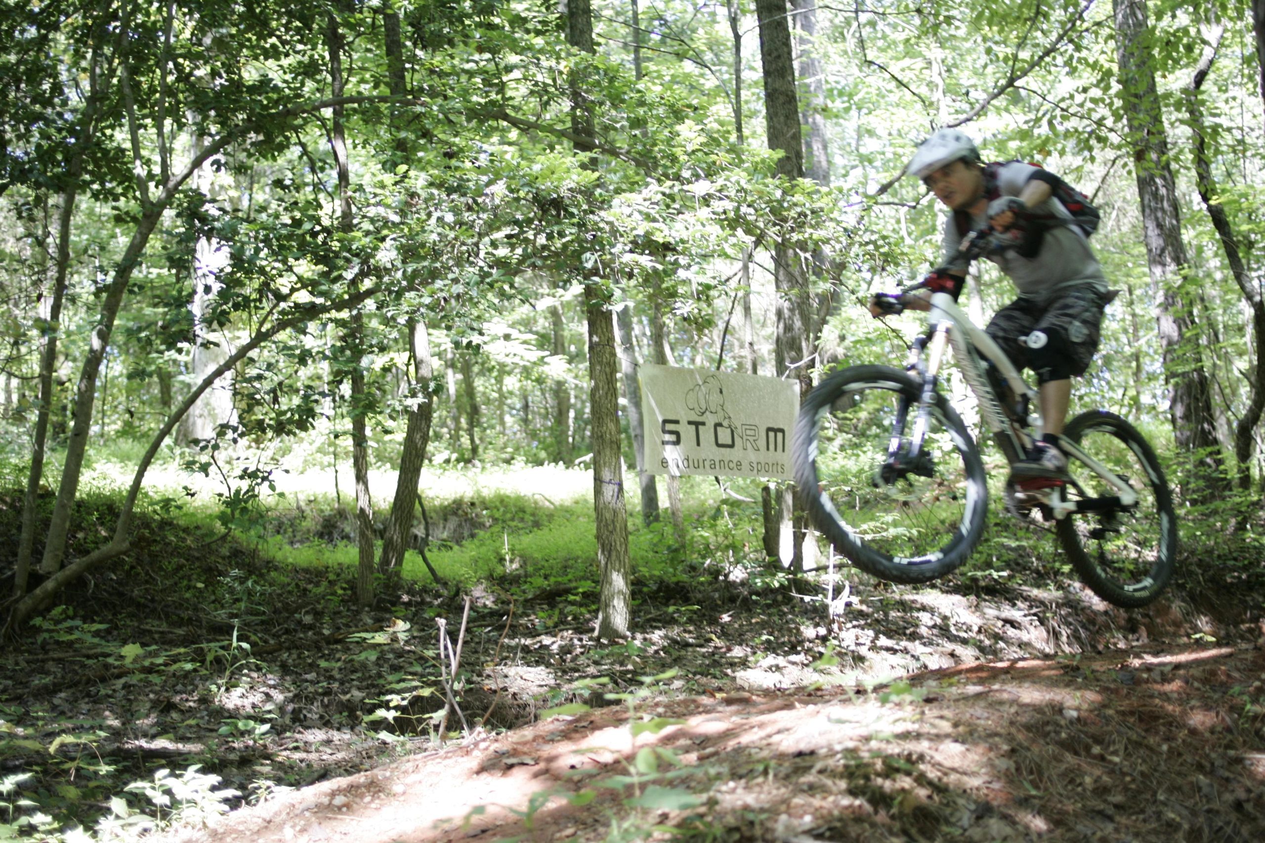 A mountain biker jumps over a dirt trail in a wooded area, with green foliage surrounding the scene. In the background, a sign marks the "STORM endurance sports" trail. San-lee Park mountain bike trail.