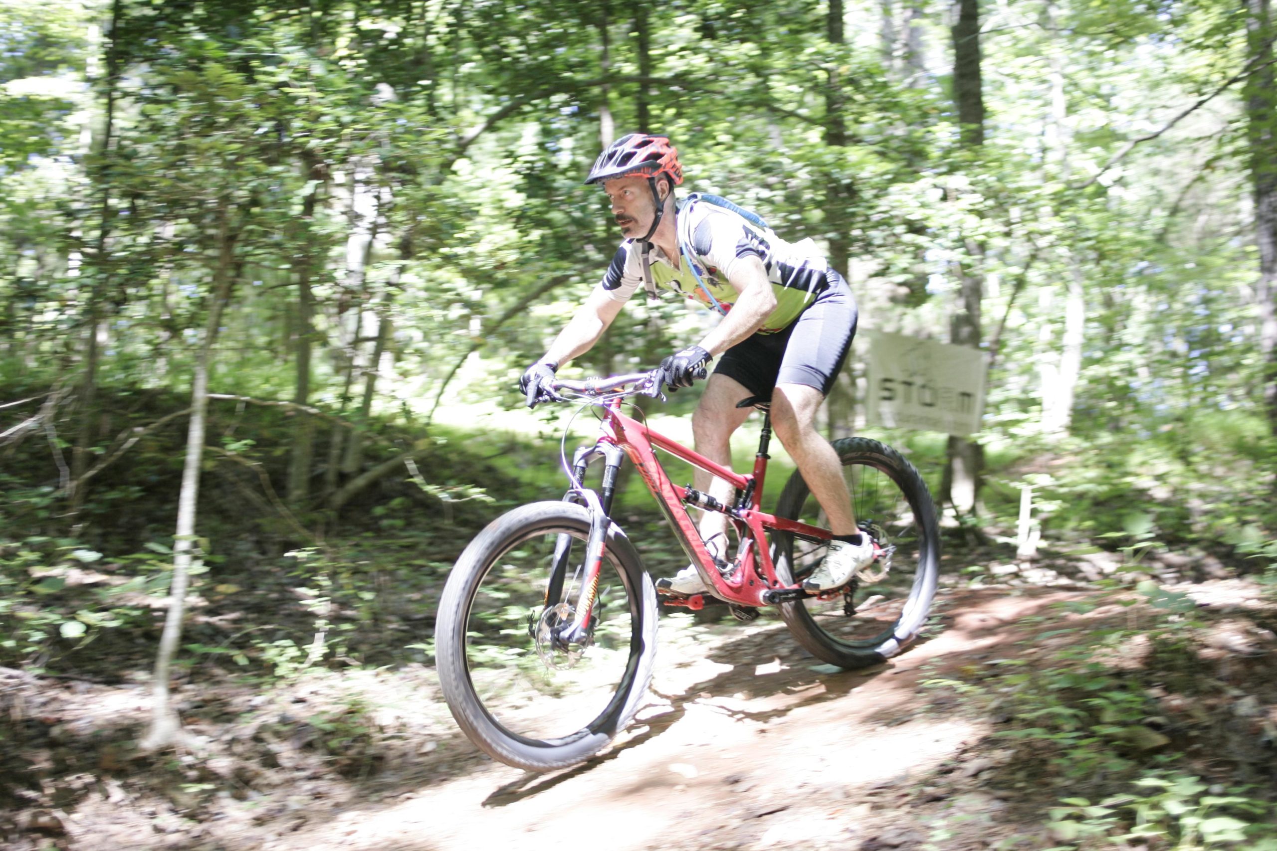 A cyclist riding a pink mountain bike down a dirt trail in a forested area, surrounded by green trees and foliage. The rider is focused and wearing a helmet, shorts, and a colorful jersey, with motion captured to convey speed. San-lee Park mountain bike trail.